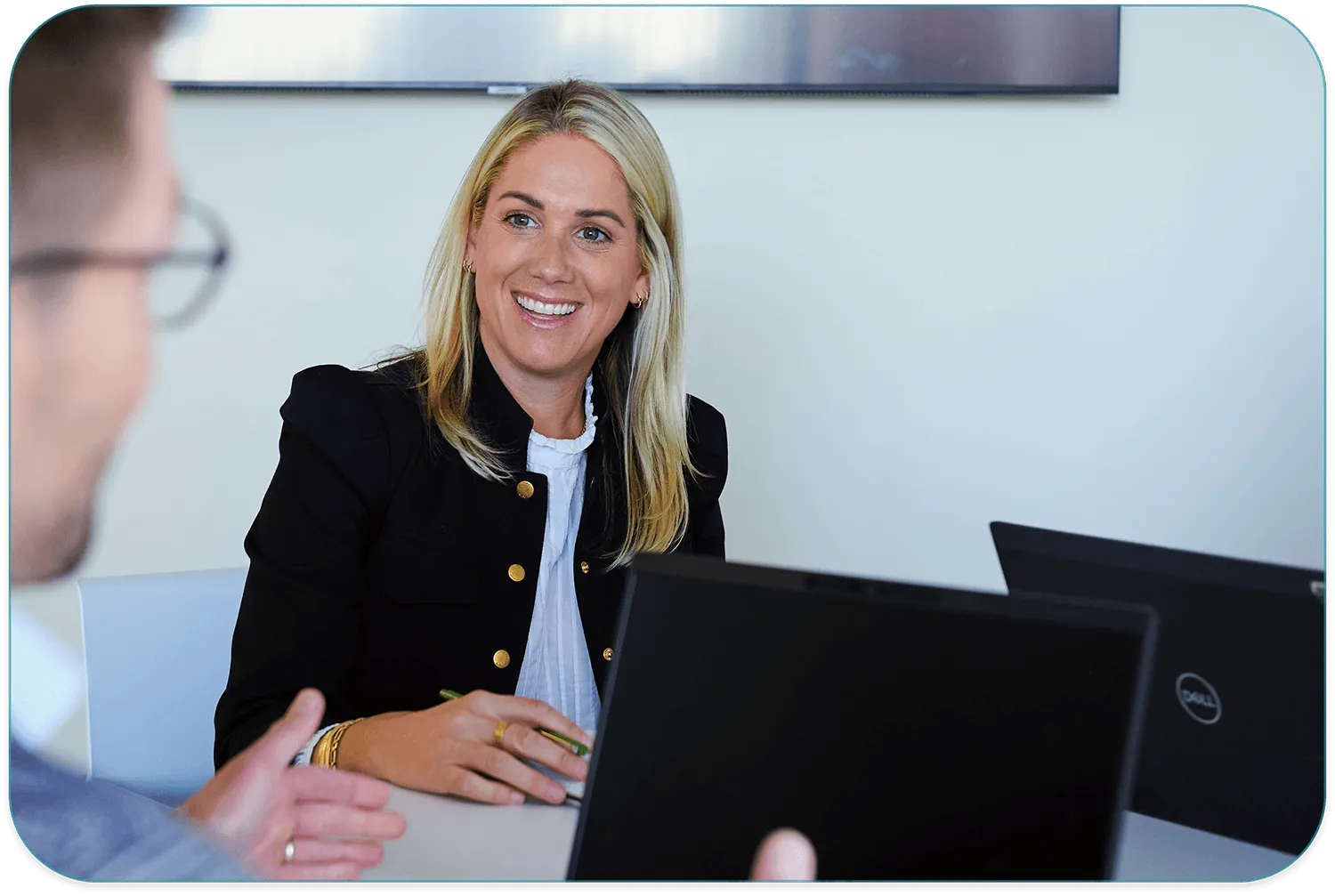 Smiling woman in a black blazer sitting at a table across from a colleague, with laptop computers open between them in an office setting.