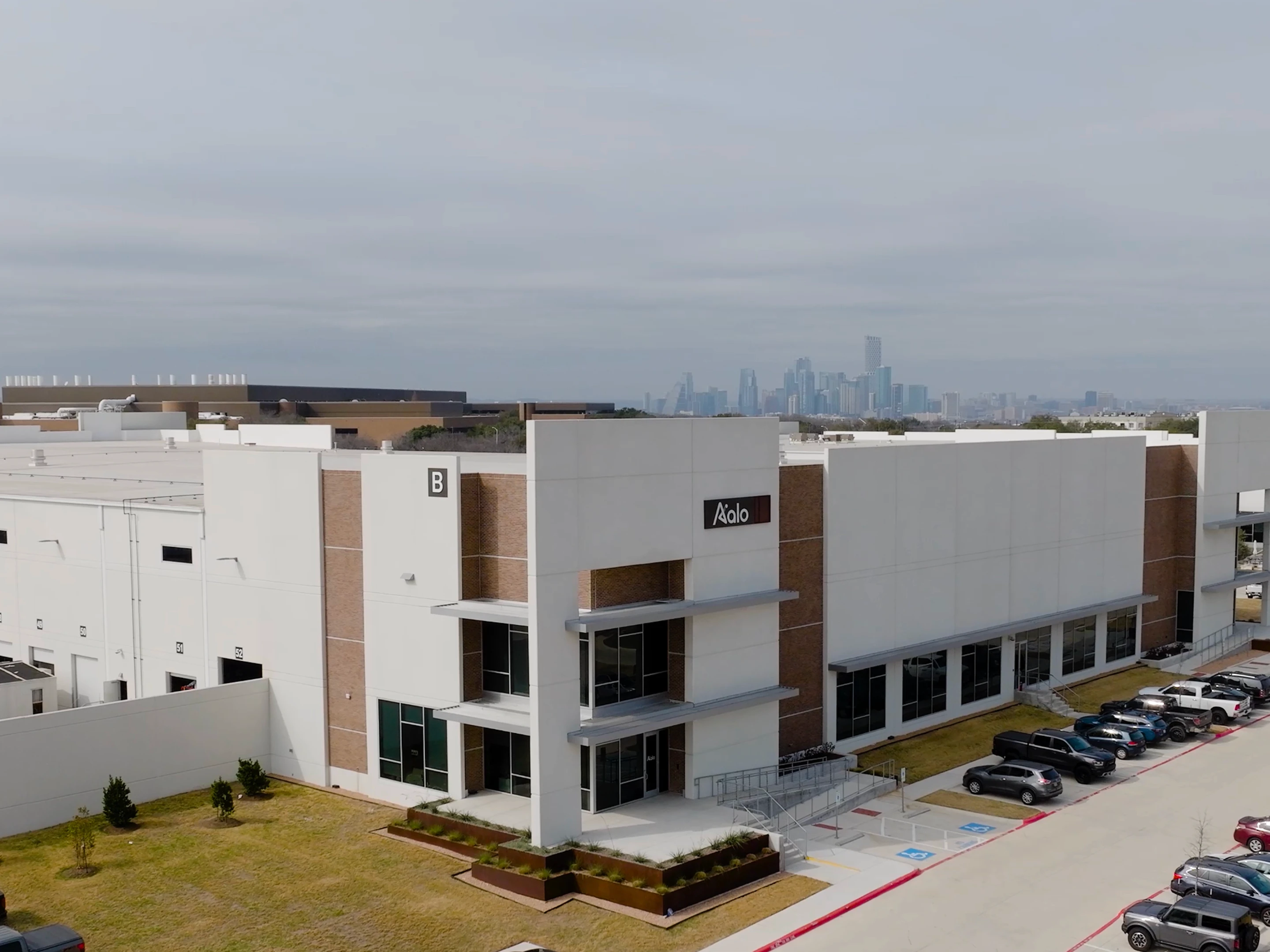 Modern commercial building with Aalo sign, parking lot, and a city skyline in the background under cloudy sky.