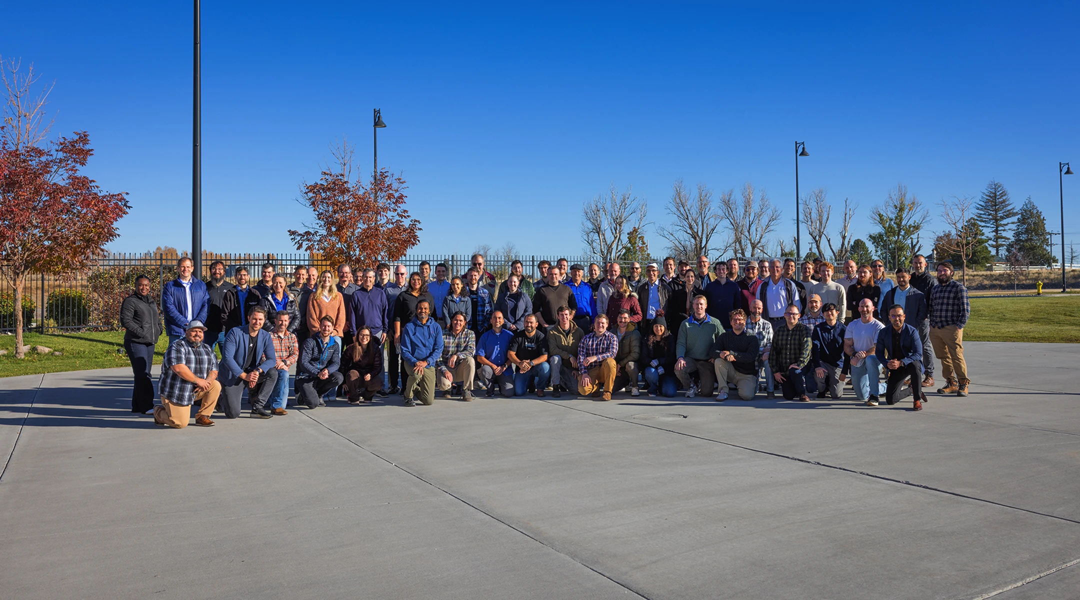 Large group of people posing outdoors on concrete under clear blue sky with some trees and lamp posts behind.