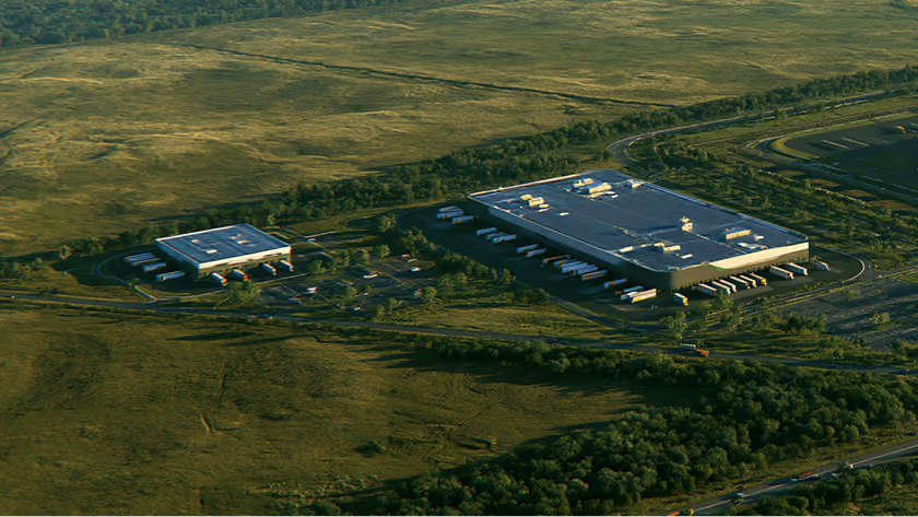 Aerial view of two warehouse buildings surrounded by green fields and trees with trucks parked outside.