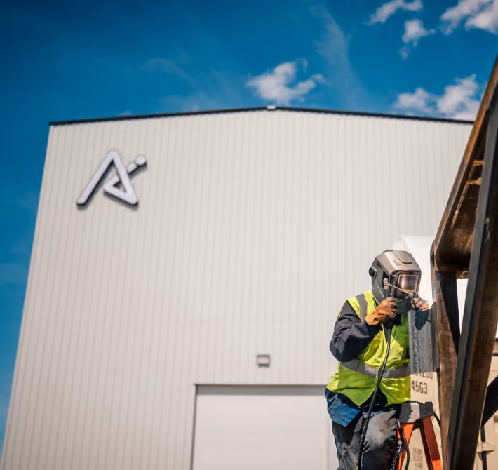 Worker in a helmet and high-visibility vest welding a metal structure outside a large building.