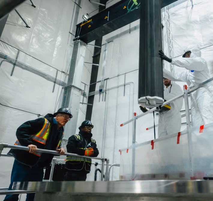 Workers in safety gear and hazmat suits operate equipment inside a large industrial facility.