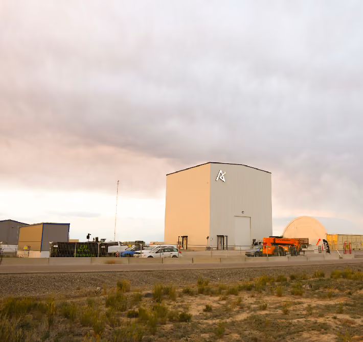 Large industrial building with logo and surrounding vehicles under a cloudy sky at sunset.