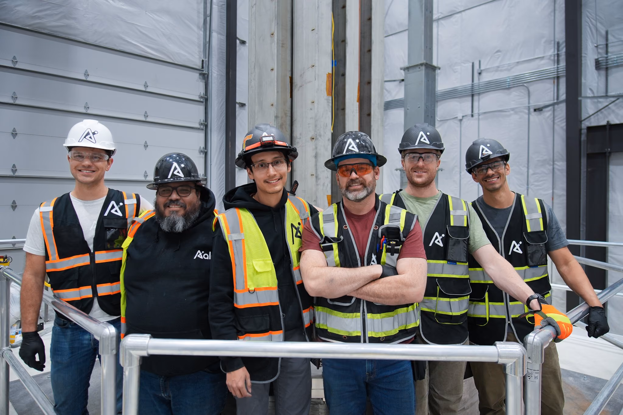Six construction workers in safety vests and helmets posing inside an industrial building.