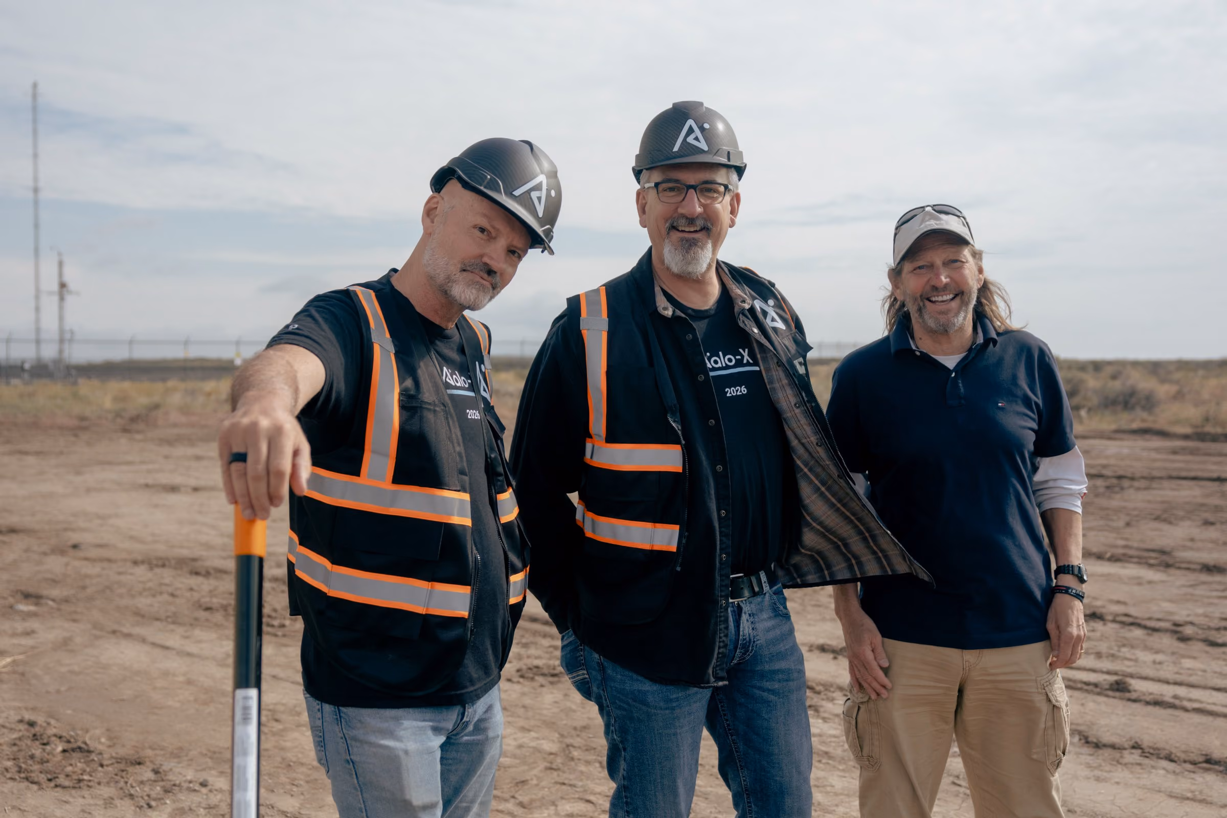 Three men smiling outdoors at a construction site, two wearing hard hats and reflective vests.