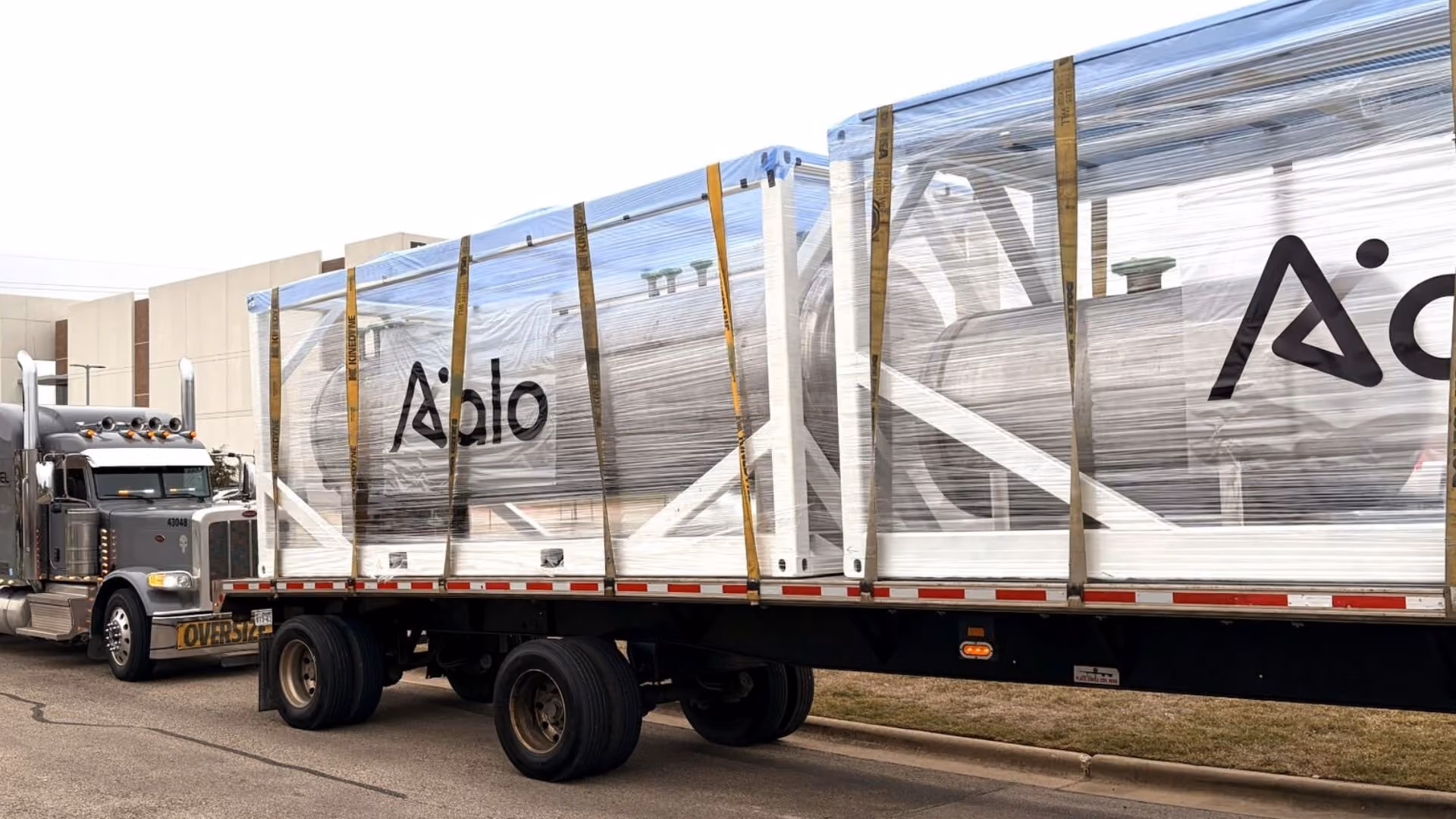 Large truck carrying a wrapped industrial reactor with Apollo logo on the side in an industrial area.
