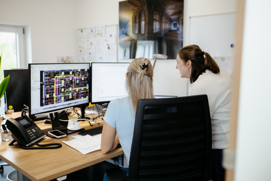 Zwei Frauen arbeiten gemeinsam an zwei Monitoren mit technischen Plänen in einem Büro.