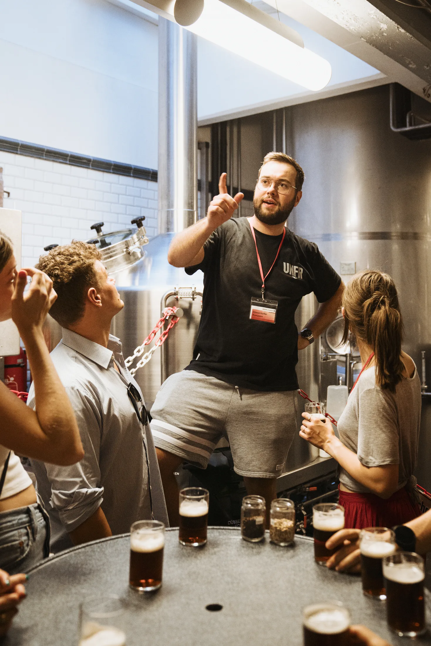 Mann erklärt Gruppe in Brauerei, umgeben von Biergläsern und Brauereigeräten.
