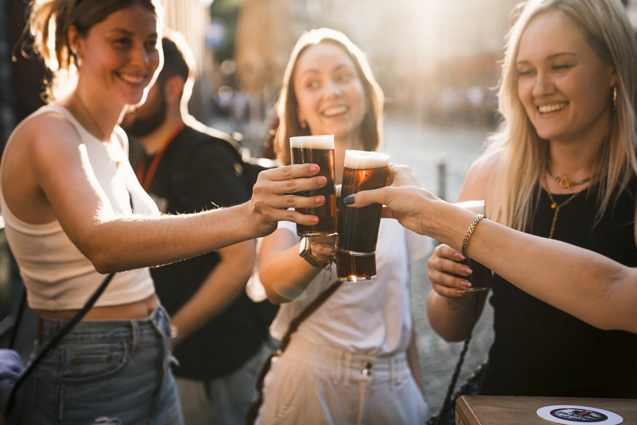 Drei lächelnde junge Frauen stoßen mit Gläsern dunklem Bier an, draußen bei sonnigem Wetter.