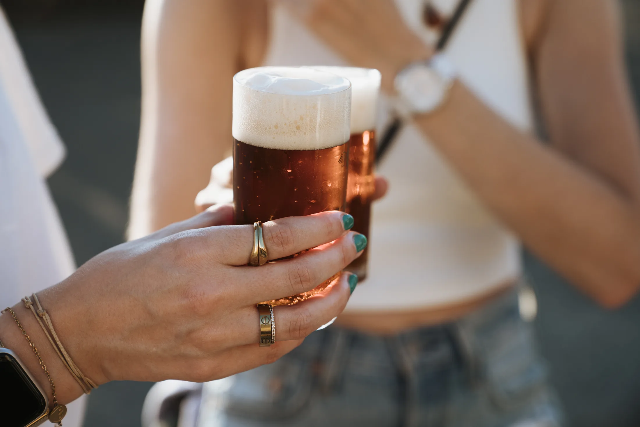 Zwei Menschen stoßen mit vollen Biergläsern an, eine Hand mit Ringen und grünen Nägeln im Vordergrund.