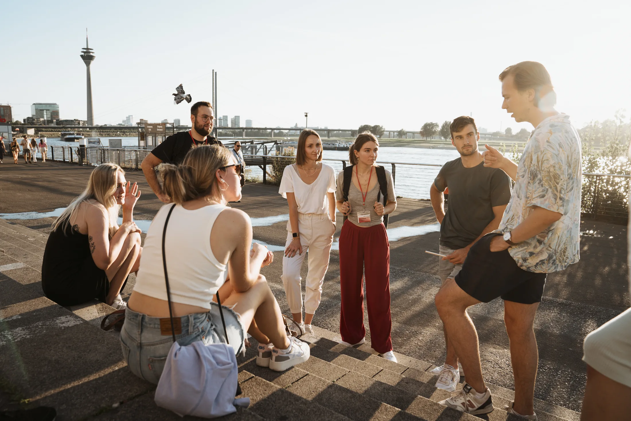 Gruppe junger Erwachsener sitzt und steht an Treppenstufen am Flussufer mit Fernsehturm im Hintergrund.