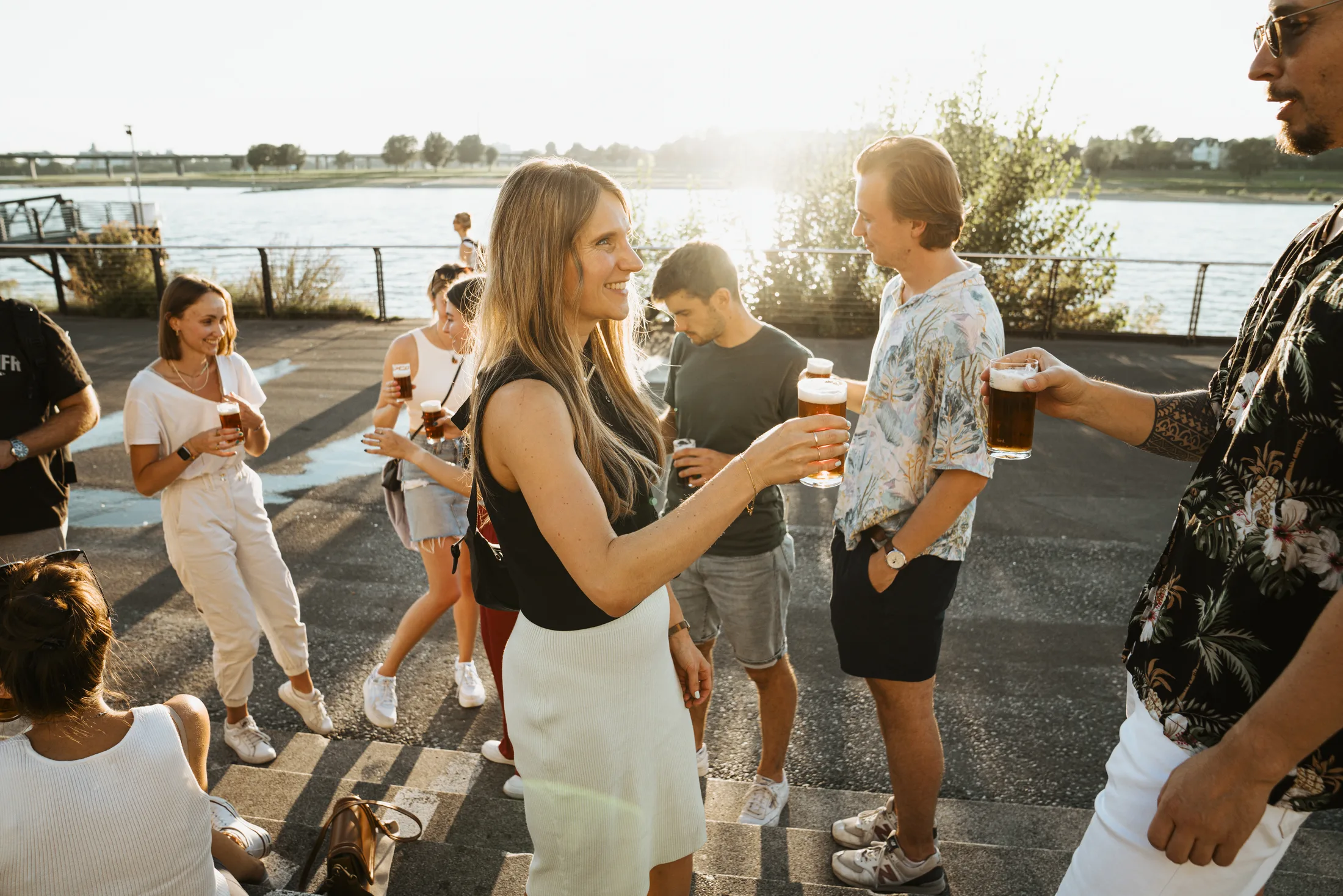 Gruppe junger Leute trinkt Bier und unterhält sich beim Treffen am Flussufer im Sonnenschein.