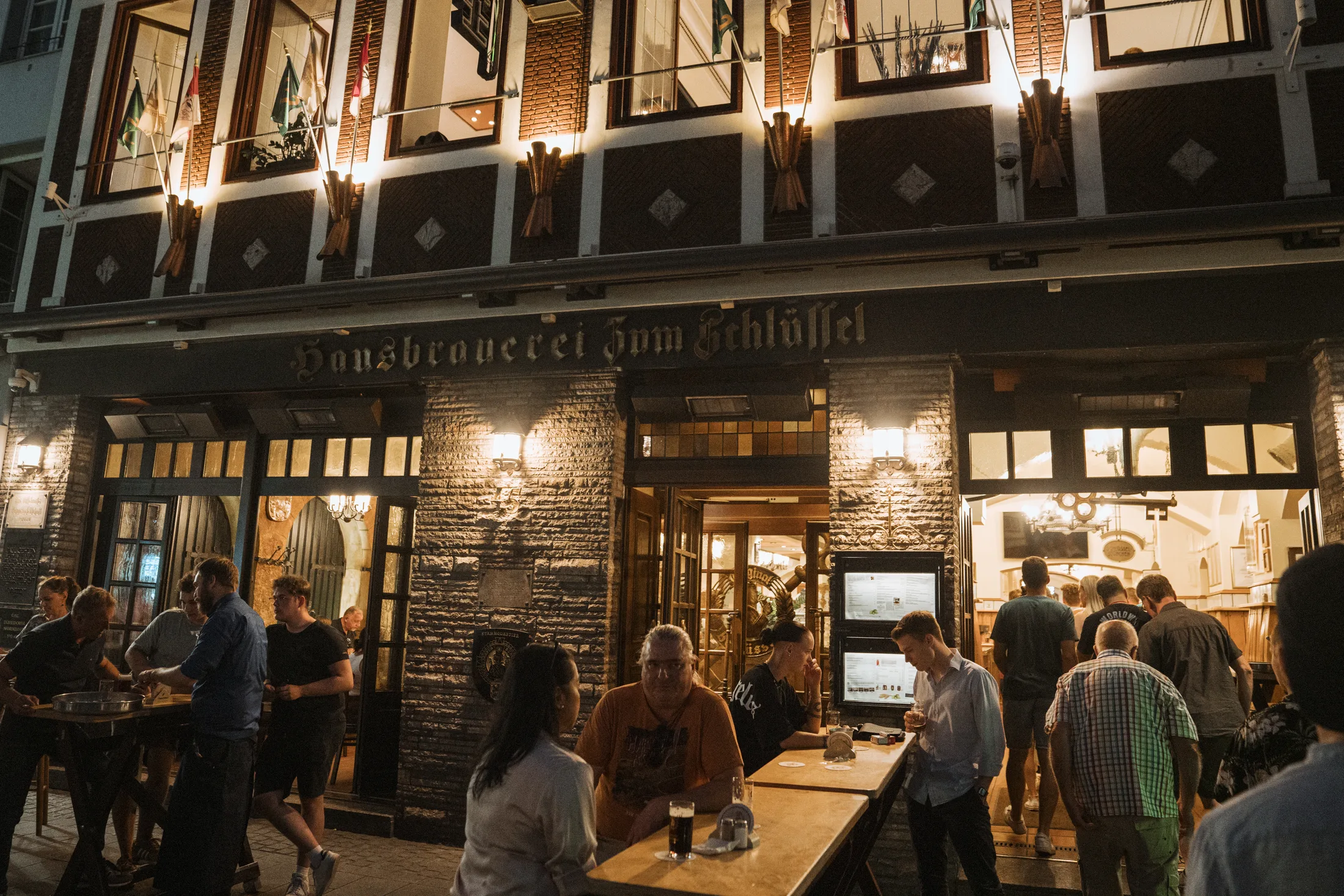 Abendaufnahme vor Hausbrauerei Zum Schlüssel mit Menschen, die draußen Tische besuchen und ins Innere blicken.