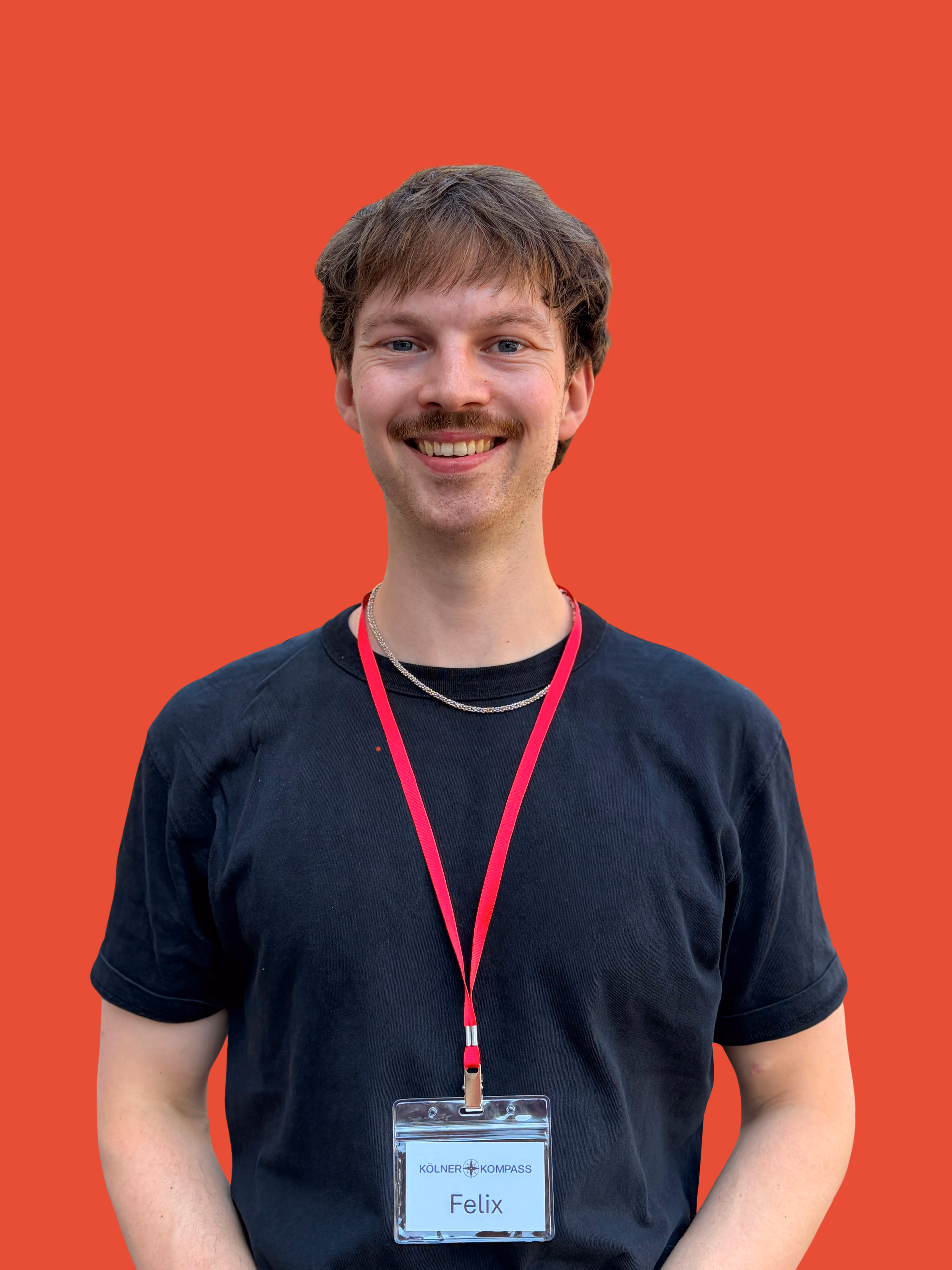 Junger Mann mit rotem Cap, schwarzem T-Shirt und Namensschild vor rotem Hintergrund.