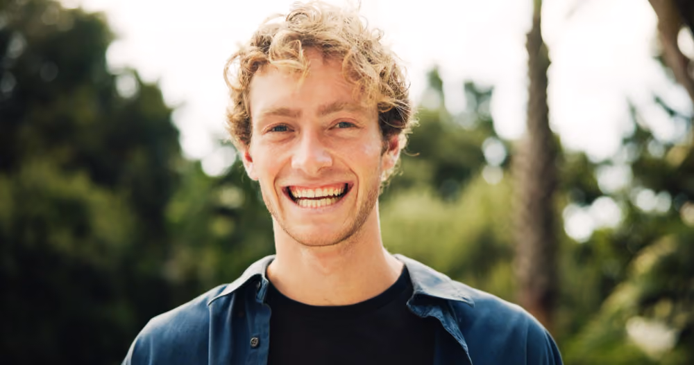 orträtfoto eines lächelnden jungen Mannes mit lockigen blonden Haaren, dunkler Jeansjacke und schwarzem Shirt, aufgenommen im Freien vor grünem Hintergrund mit Bäumen und Palme. Das Bild ist als Tablet Mockup auf blauem Gradient Hintergrund dargestellt.
