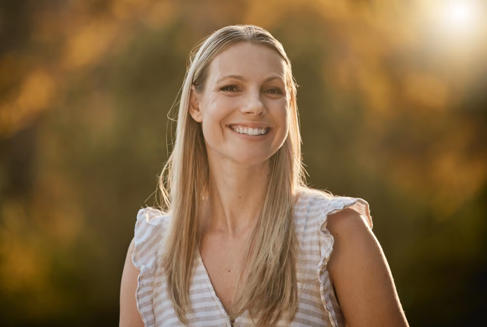 Porträtfoto einer lächelnden blonden Frau mit gestreiftem Sommerkleid, aufgenommen im Freien im warmen Abendlicht mit unscharfem grün goldenem Hintergrund. Das Bild ist als Tablet Mockup auf blauem Gradient Hintergrund dargestellt.