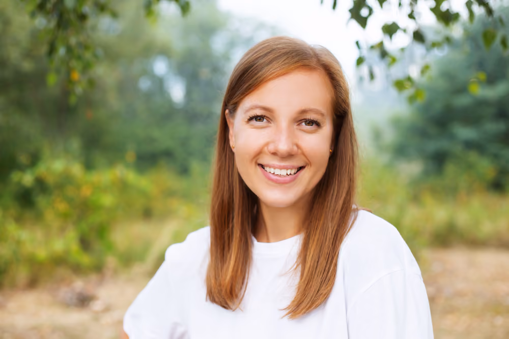 Porträtfoto einer lächelnden Frau mit langen rötlich blonden Haaren in weißem Shirt, aufgenommen im Freien vor grünem Hintergrund mit Bäumen. Das Bild ist als Tablet Mockup auf blauem Gradient Hintergrund dargestellt.