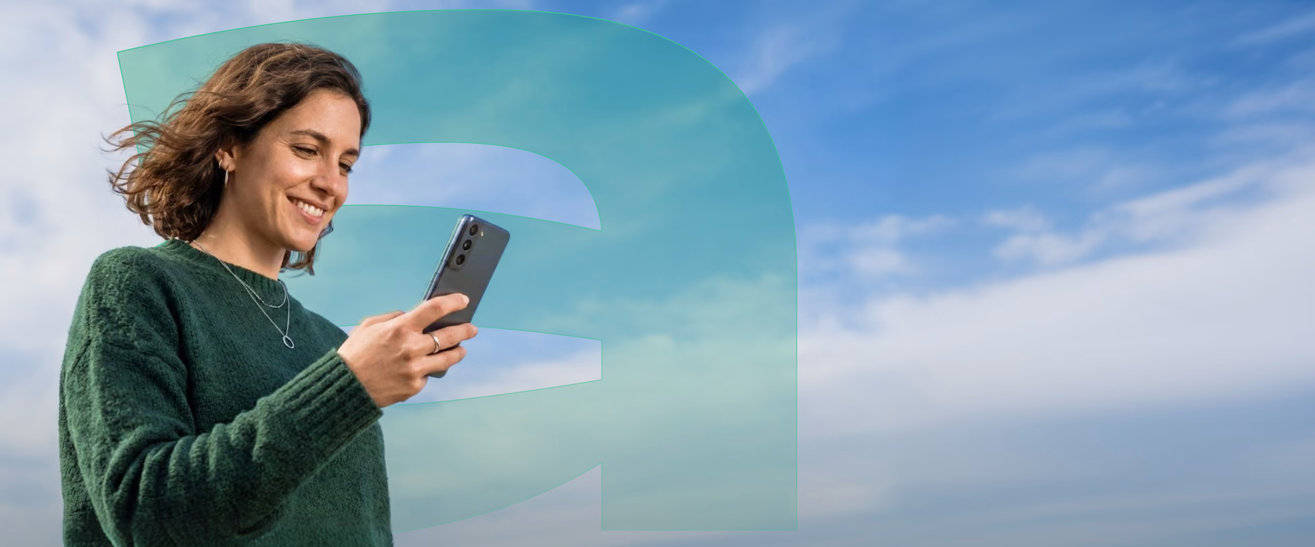 A smiling young woman with curly hair wearing a green sweater looks at her smartphone against a blue sky with some clouds.
