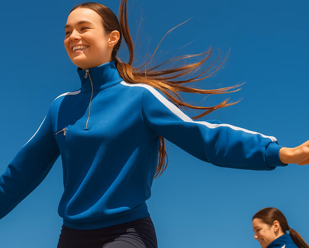 Smiling woman with long brown hair in a blue zip-up jacket with white stripes on sleeves, arms outstretched against a clear blue sky, with another woman in a similar jacket in the background.