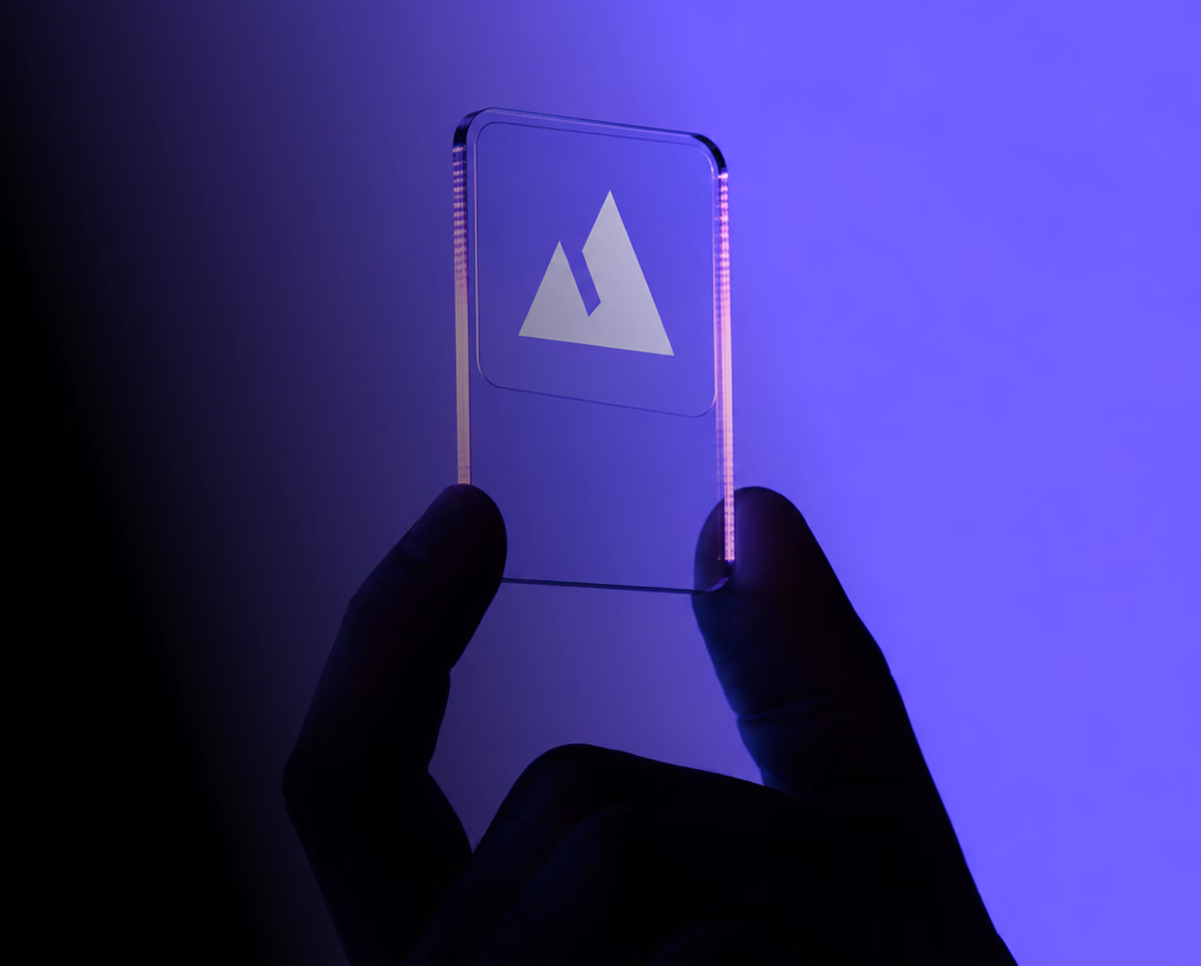 Hand holding a clear acrylic card engraved with a white mountain logo, lit against a bright violet background