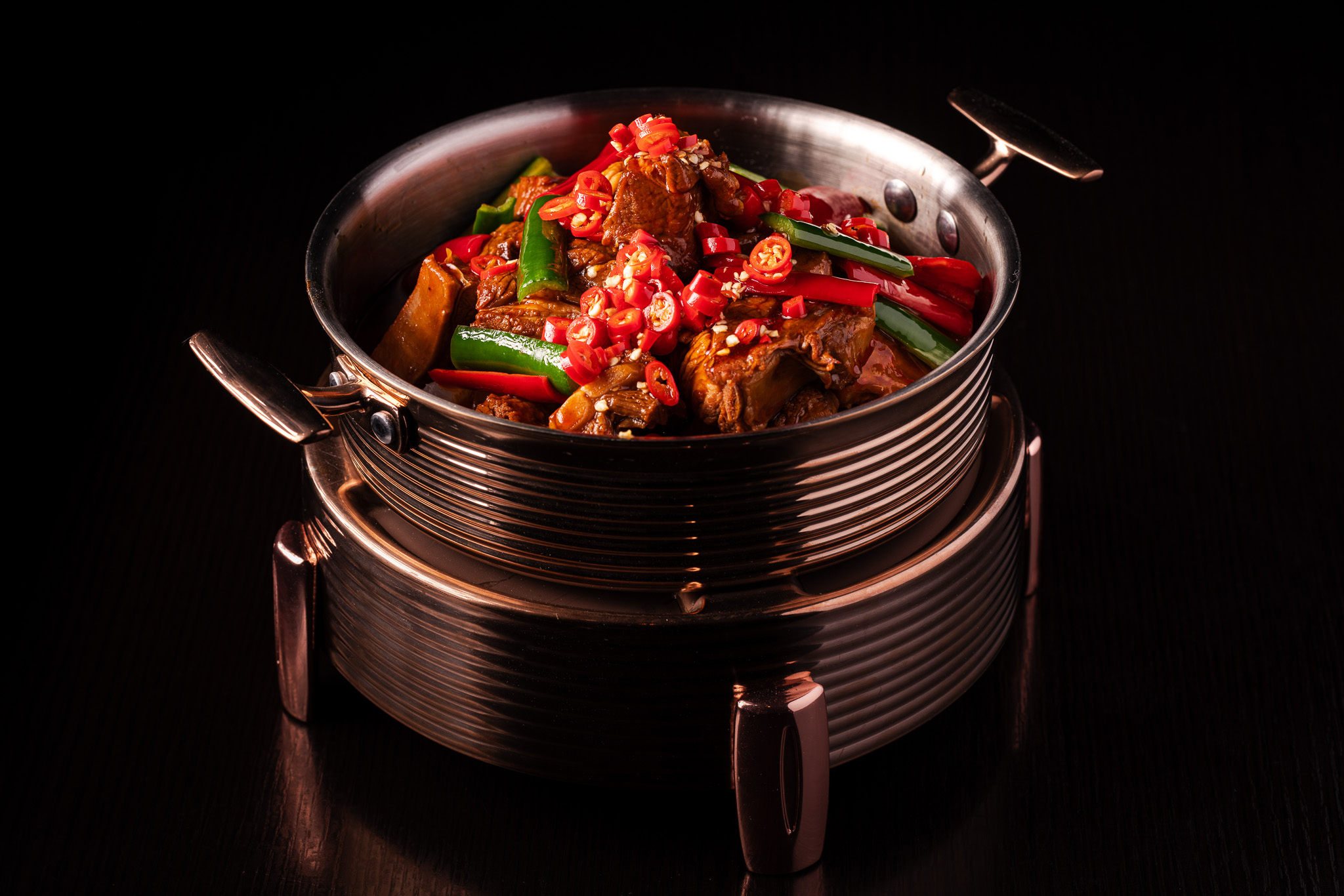 Metal bowl with braised meat dish garnished with sliced red and green chili peppers on a matching metallic stand, against a dark background.