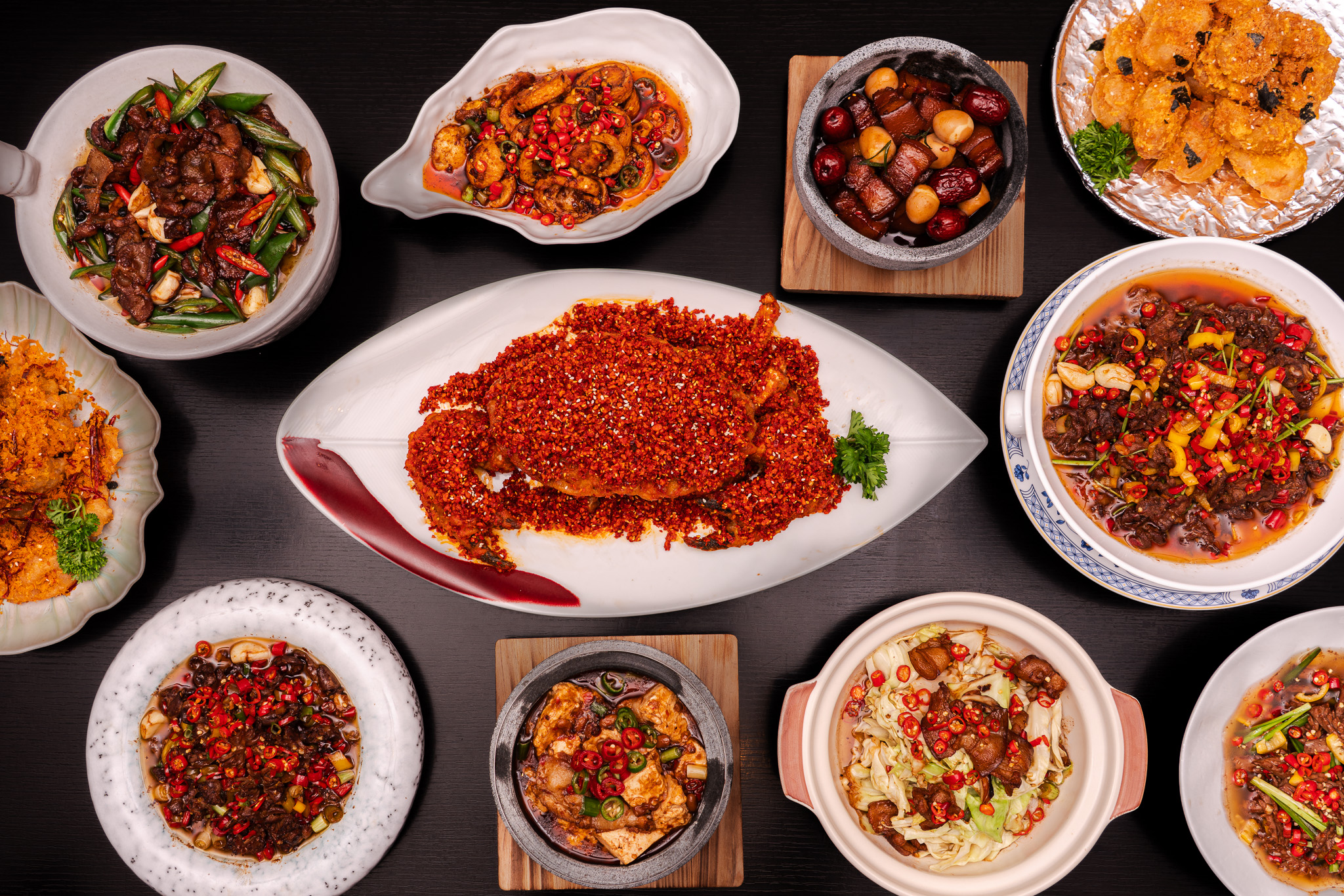 Overhead view of a variety of Chinese dishes on a dark table including a spicy crab covered in red chili flakes on a large white plate, multiple bowls of stir-fried meats and vegetables with chili peppers, tofu, mushrooms, and crispy fried items garnished with fresh herbs.
