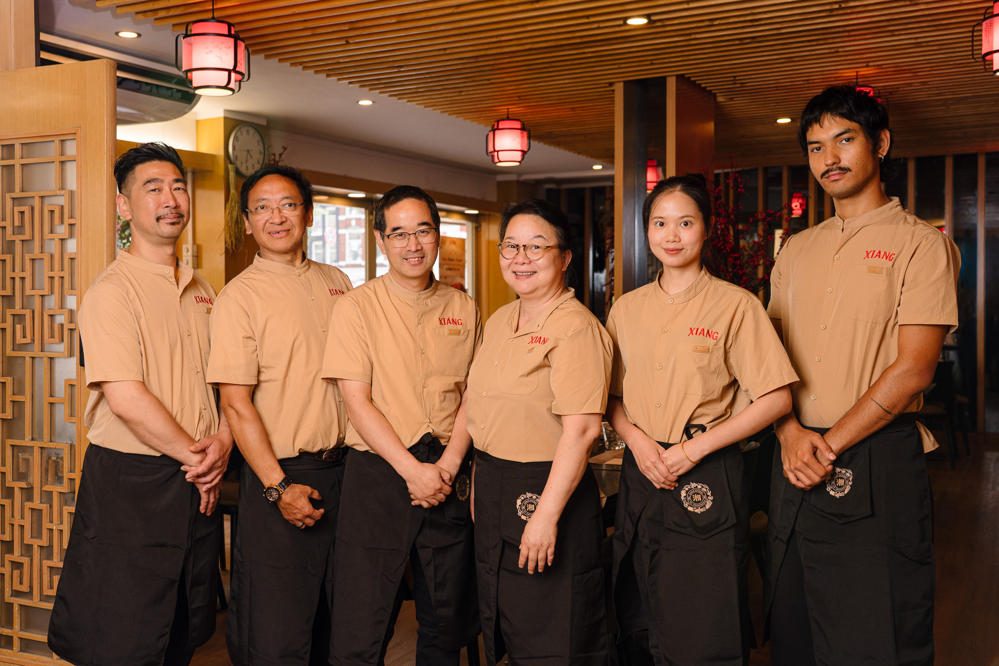 Six restaurant staff members standing side by side inside a warmly lit restaurant, wearing tan shirts with the word 'XIANG' and black aprons, smiling and facing the camera.