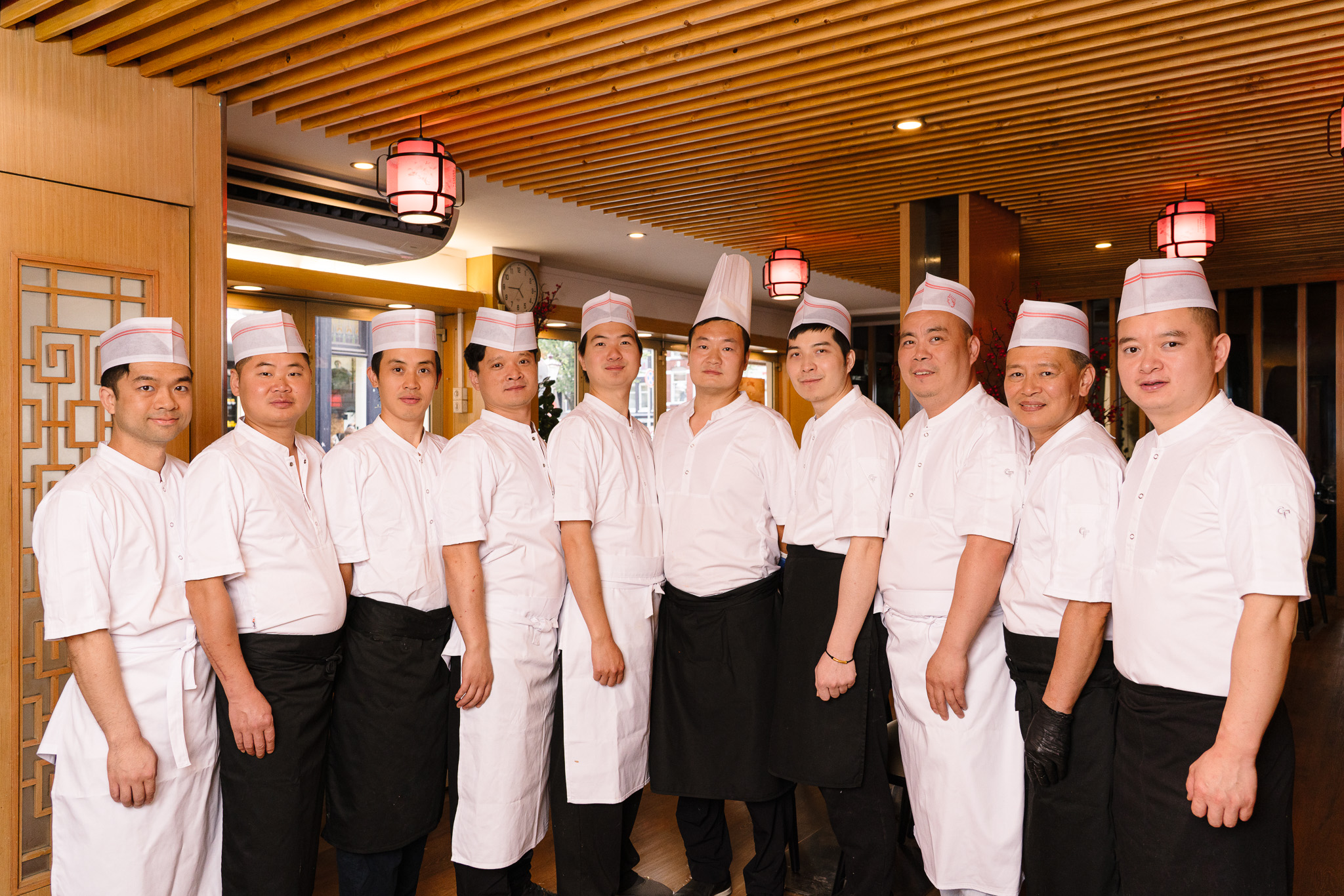 Group of eleven male chefs in white uniforms and hats standing side by side inside a restaurant with wooden decor and hanging lanterns.