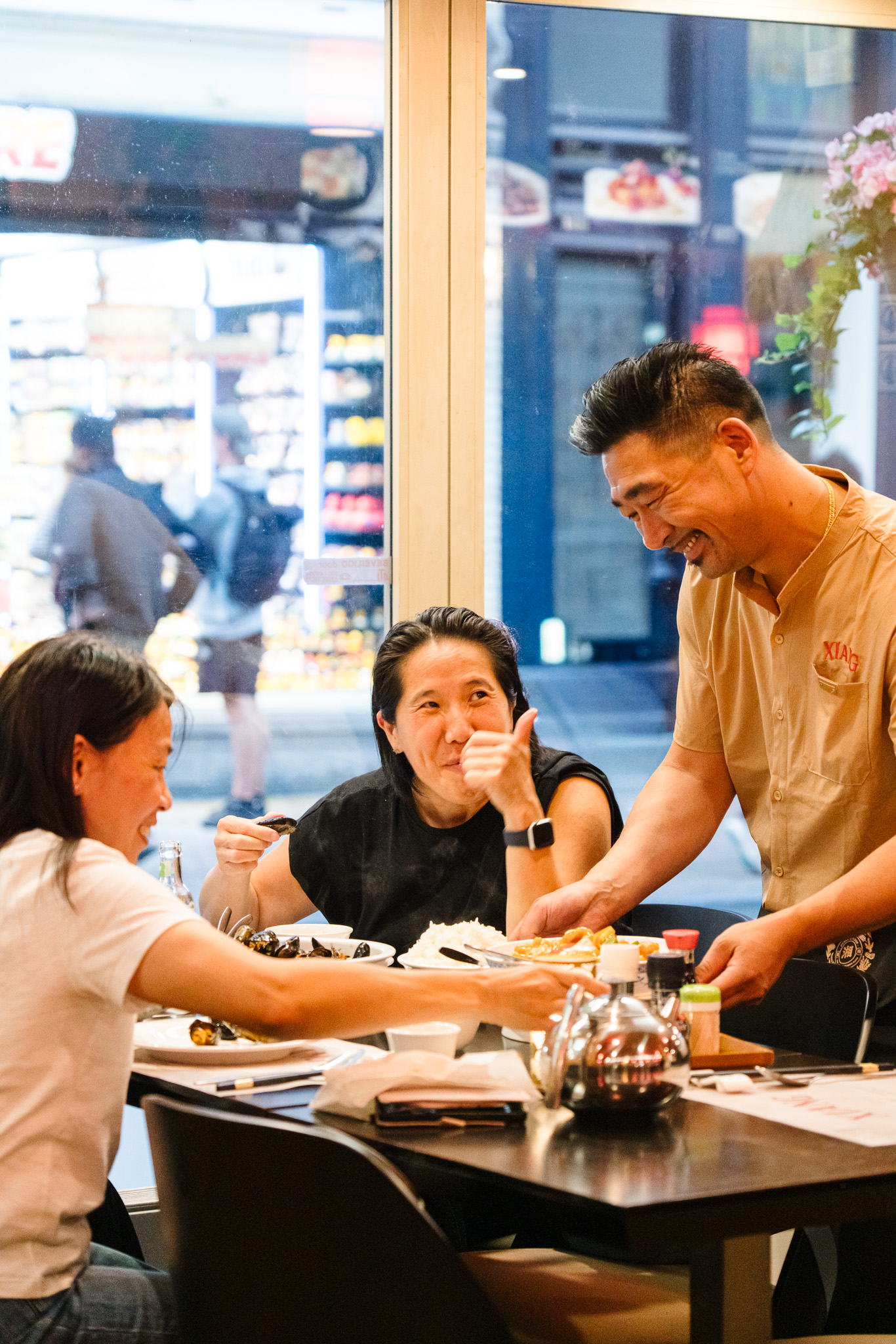 Two women seated at a restaurant table eating while a smiling male server places a dish in front of them near a window showing a blurred street scene outside.