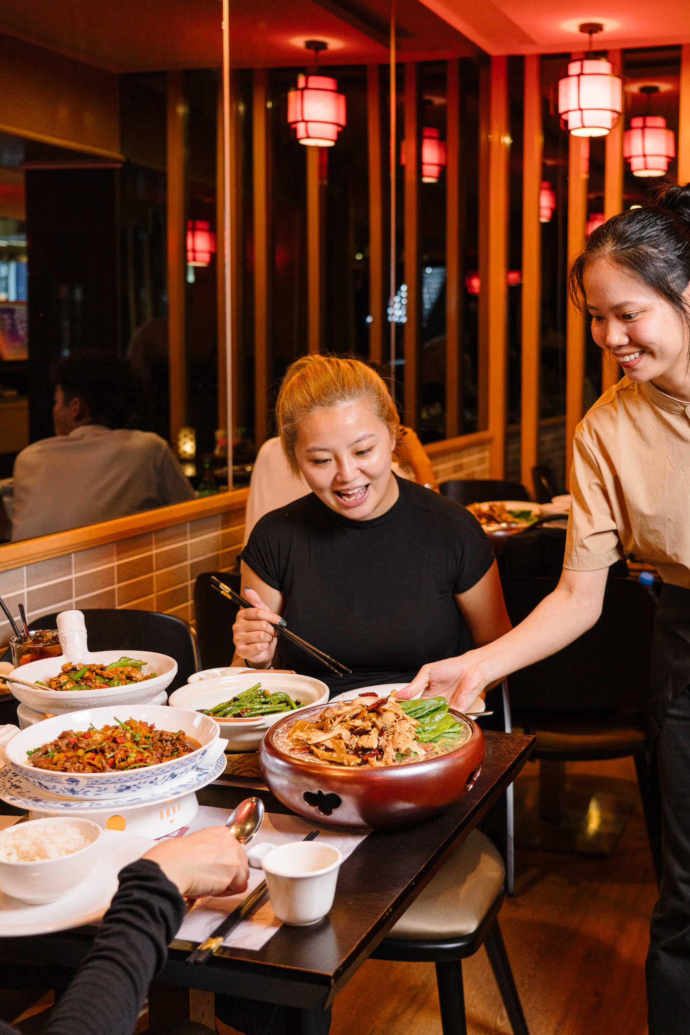 A woman with light brown hair wearing a black shirt sits at a restaurant table filled with various Asian dishes including green beans, a large bowl of meat stew, and a bowl of rice. A smiling waitress in a beige shirt serves the meat stew to the woman. The background has wooden paneling and red ceiling lights reflected in mirrors.
