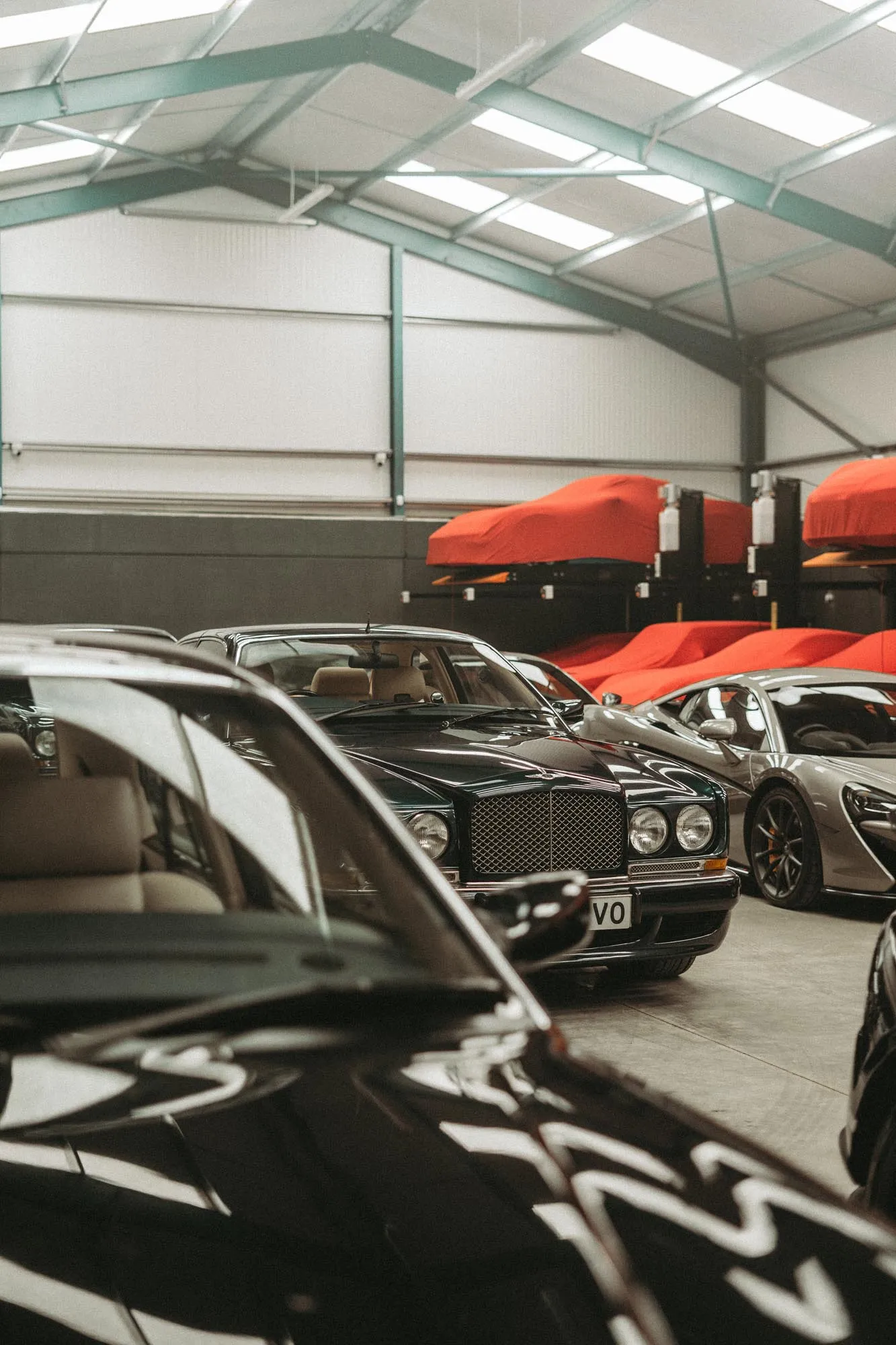 Indoor car storage with luxury cars, including a classic Bentley in the center and sports cars nearby, with several cars covered in red protective covers.