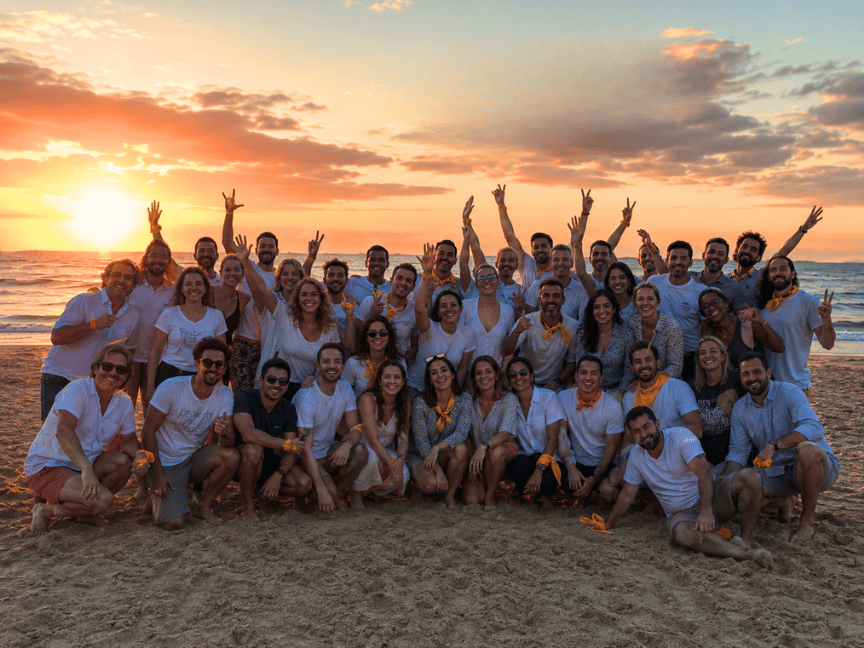 Groupe d'environ quarante personnes souriantes posant sur une plage au coucher du soleil avec des foulards orange autour du cou ou au poignet, certaines faisant des gestes de paix ou levant les bras.