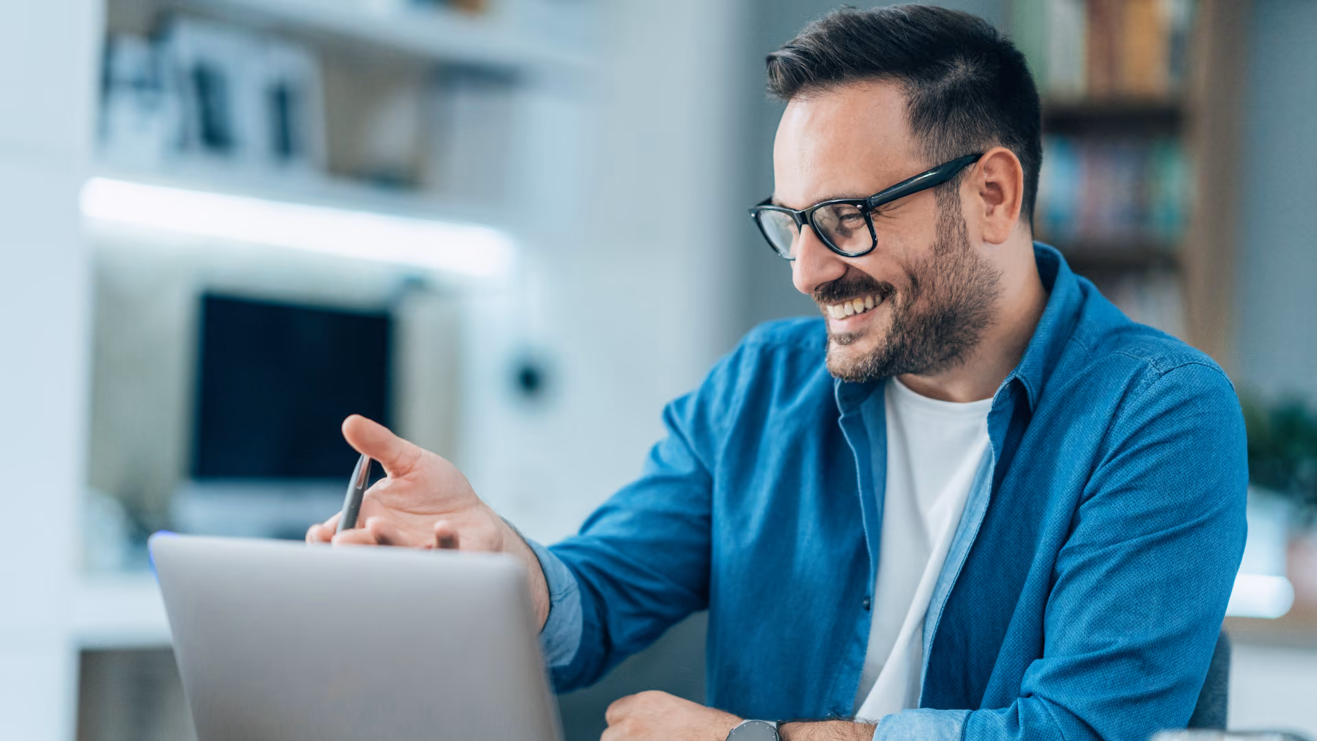 Un homme souriant portant des lunettes et une chemise bleue, discutant devant un ordinateur portable dans un intérieur lumineux.