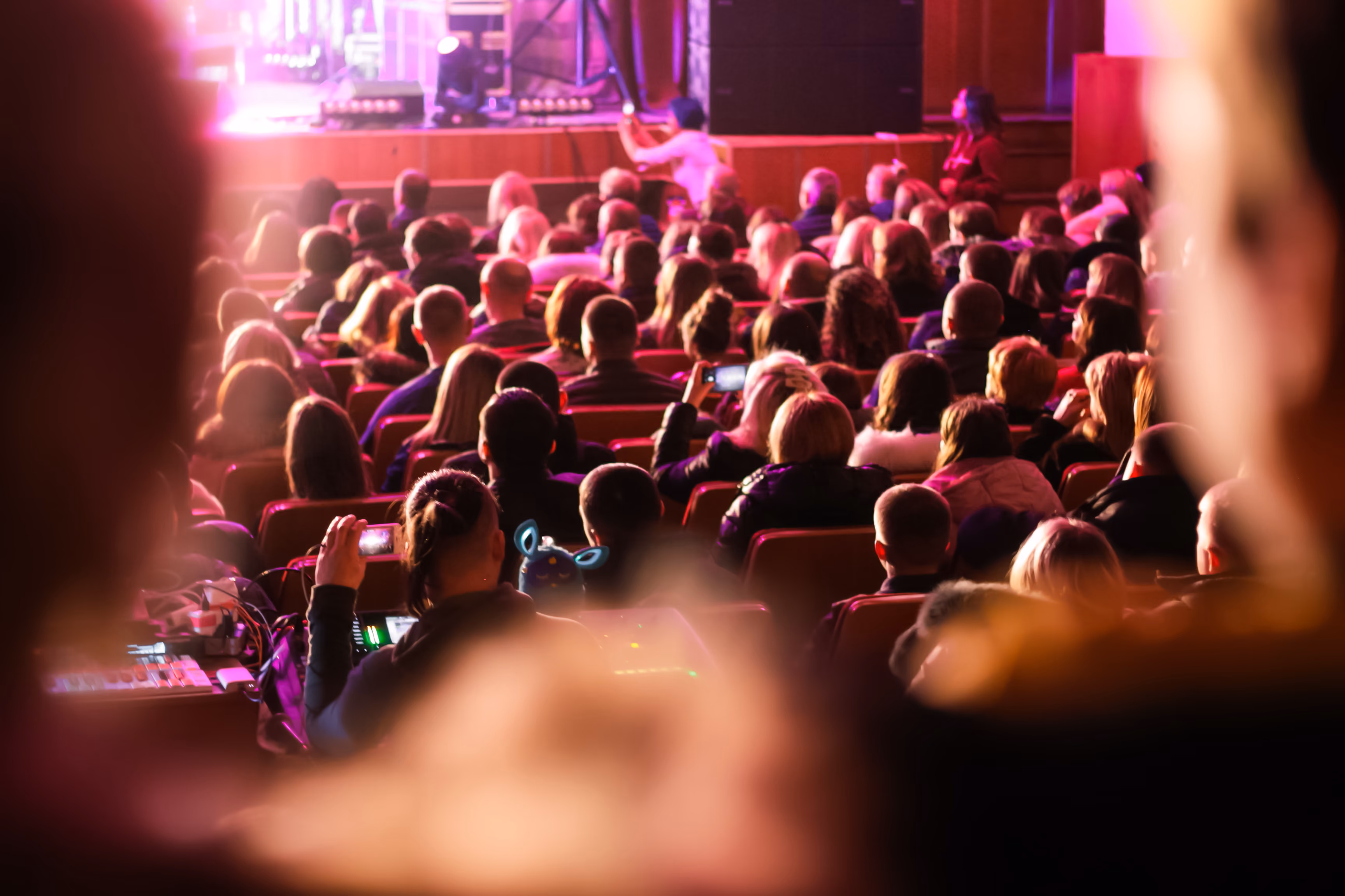 Public nombreux assis dans une salle de spectacle, certains prennent des photos ou vidéos avec leurs téléphones, lumière rosée éclaire la scène devant eux.