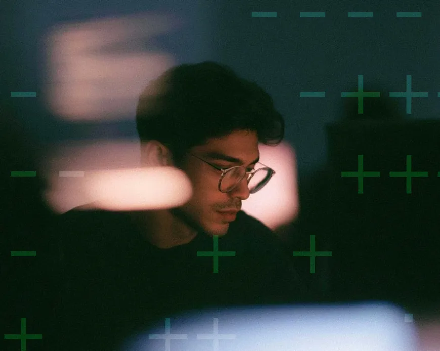 Woman pointing at computer screen with code, sitting at desk with keyboard and headphones.