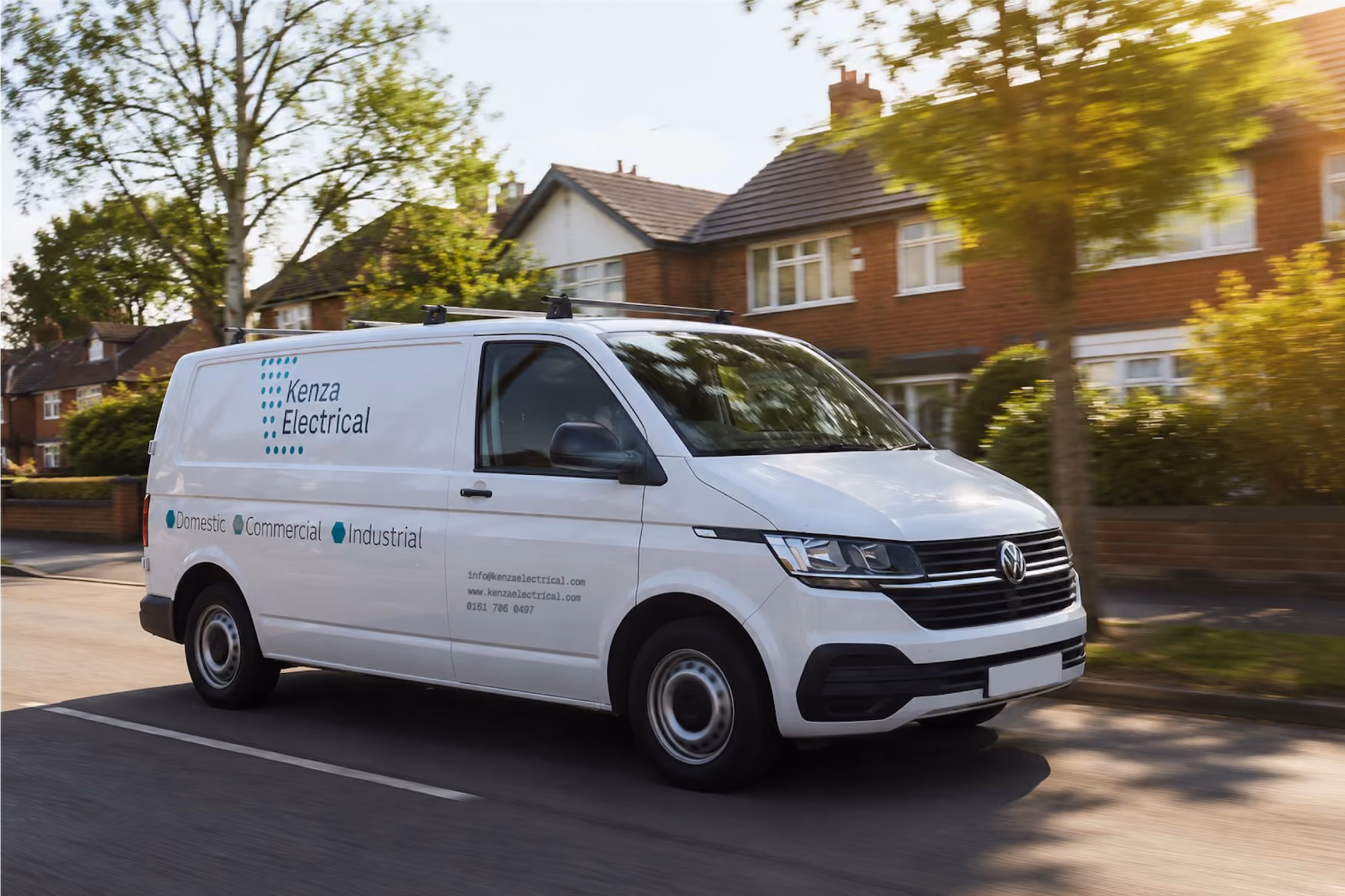 White Kenza Electrical van driving on a suburban street with houses and trees in the background.