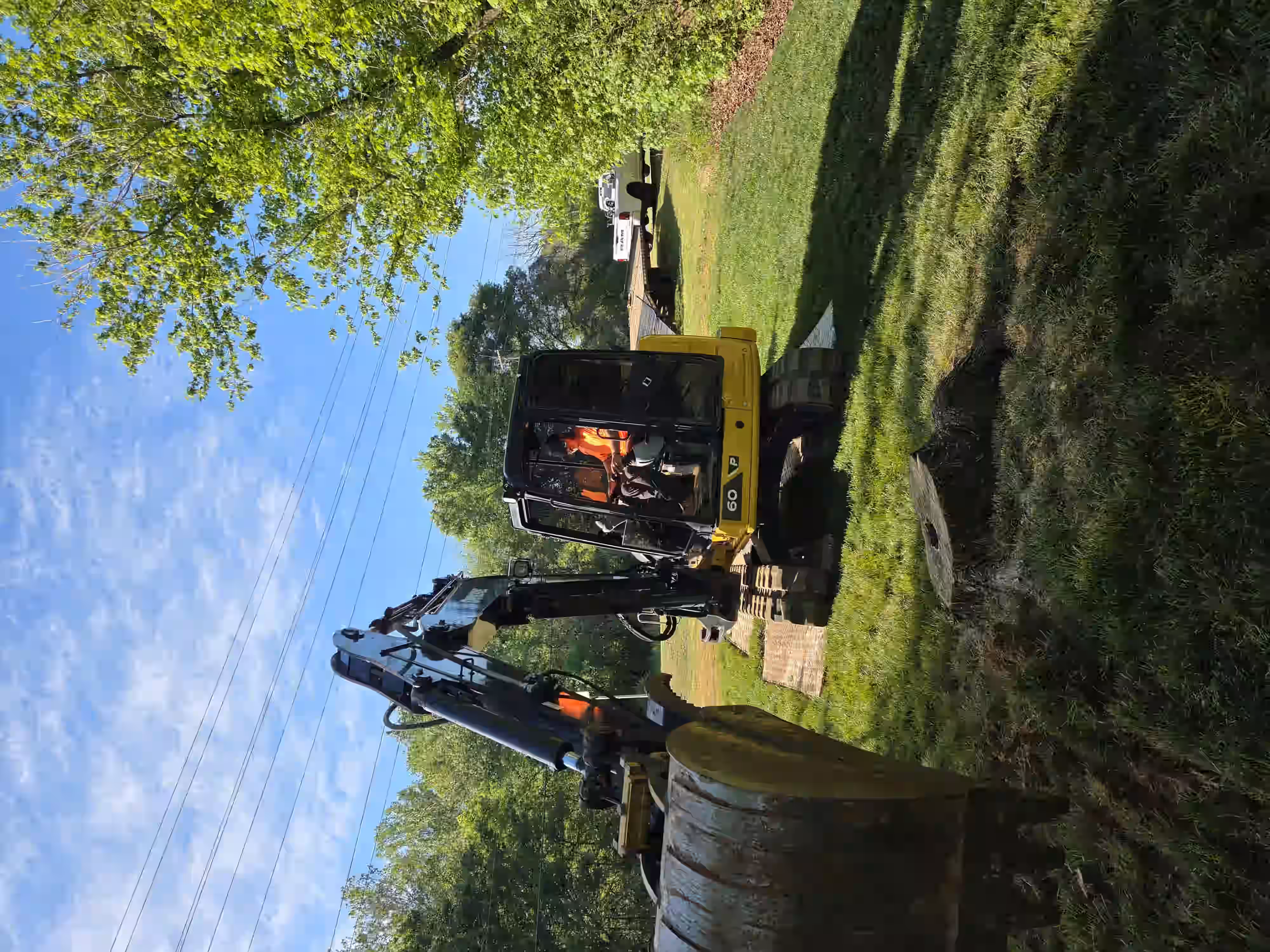 Worker in orange shirt operates a small excavator on grassy yard near tree stump and white truck.