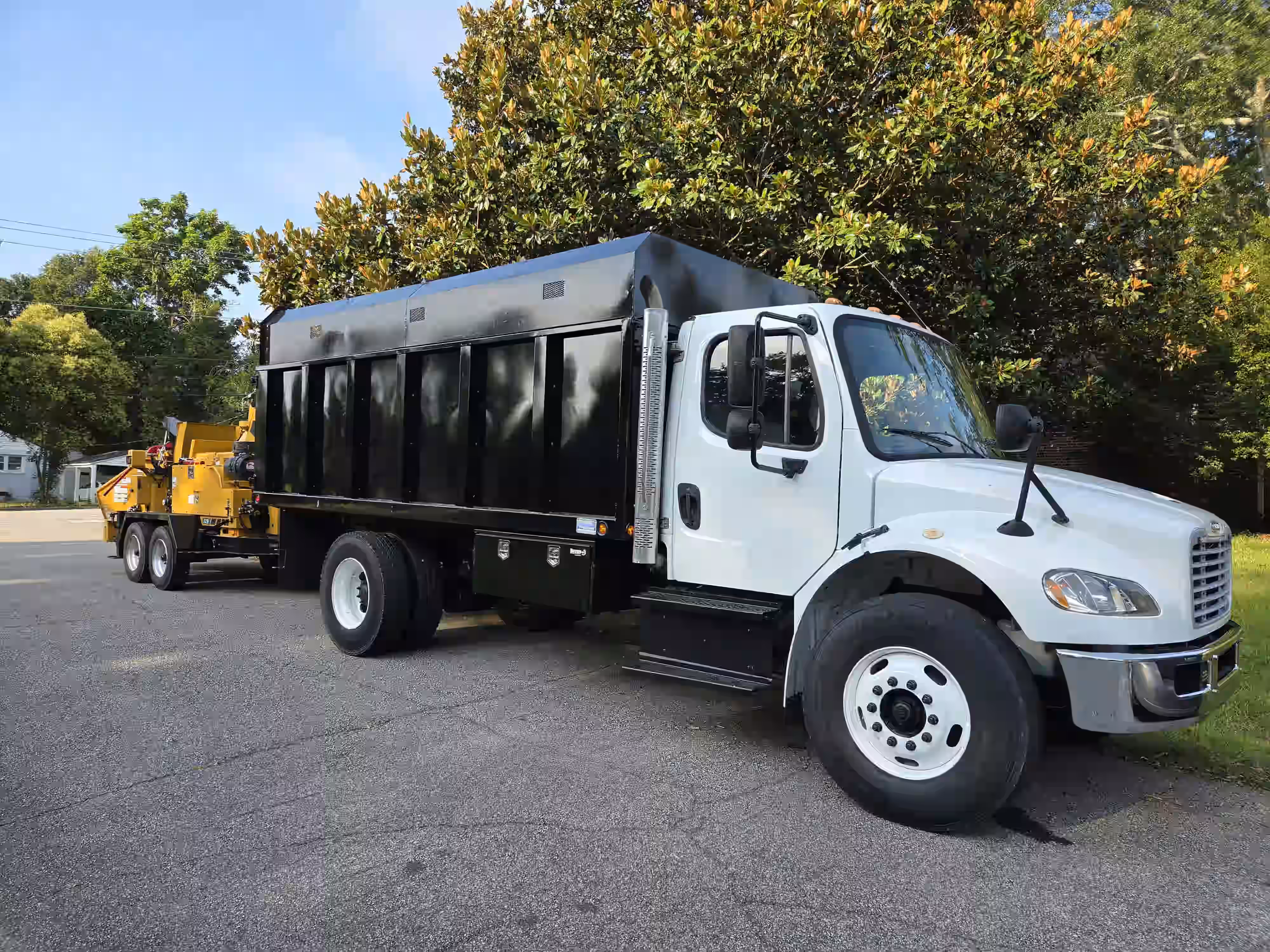 White and black dump truck parked on pavement with trees and a yellow wood chipper trailer attached.