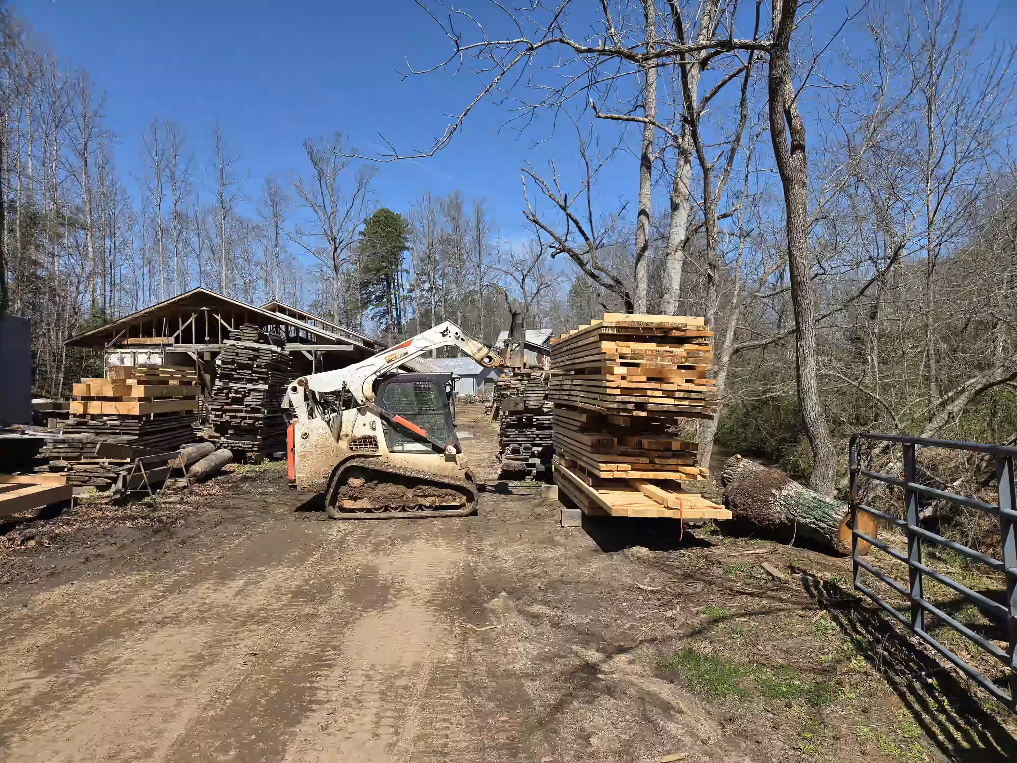 Construction vehicle with tracked wheels lifting stacked lumber at a wood yard under clear blue sky.