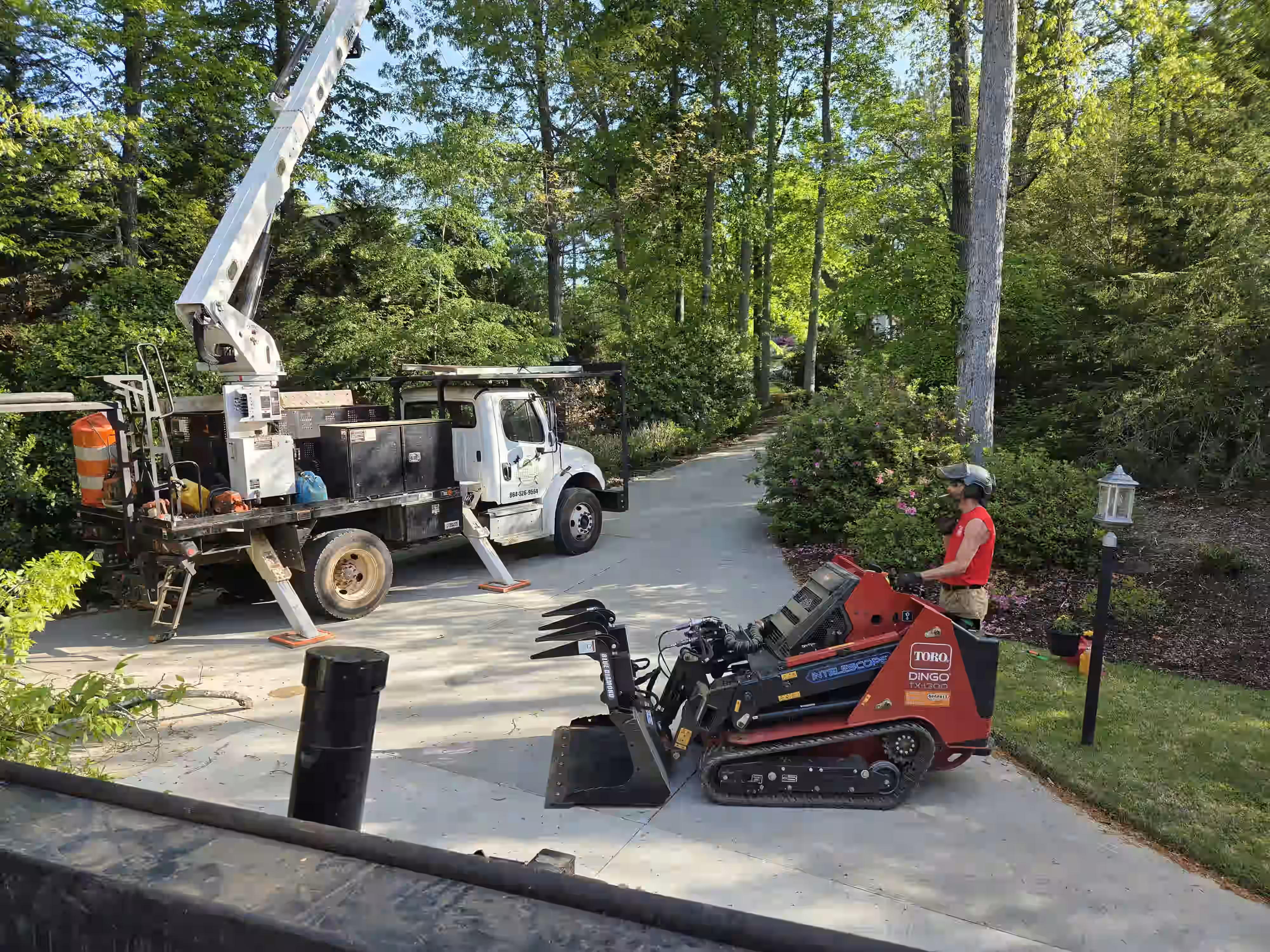 Person operating a red Toro Dingo compact loader on driveway near a bucket lift truck and green trees.