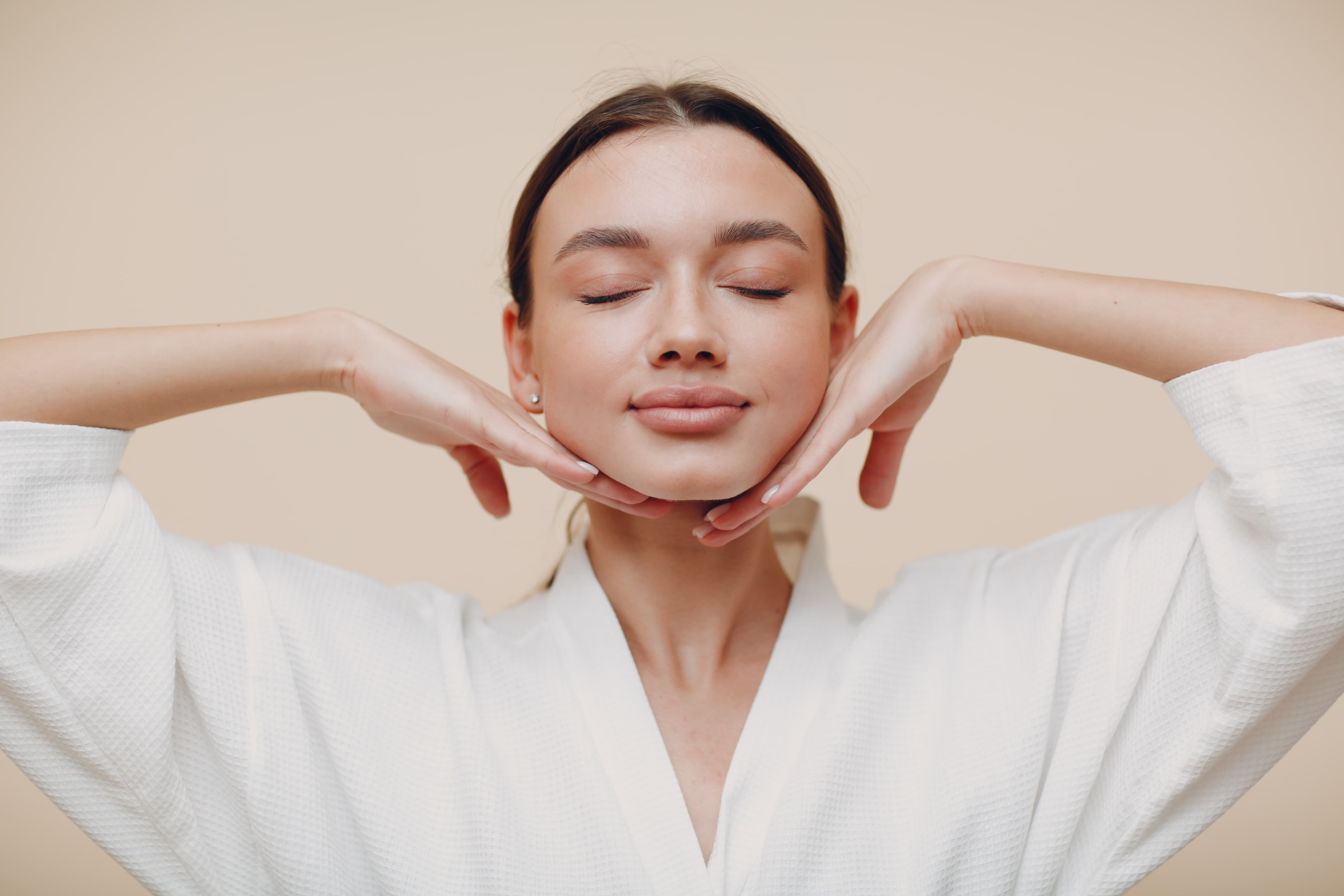 Woman in white robe with eyes closed gently holding her face with both hands near jawline.