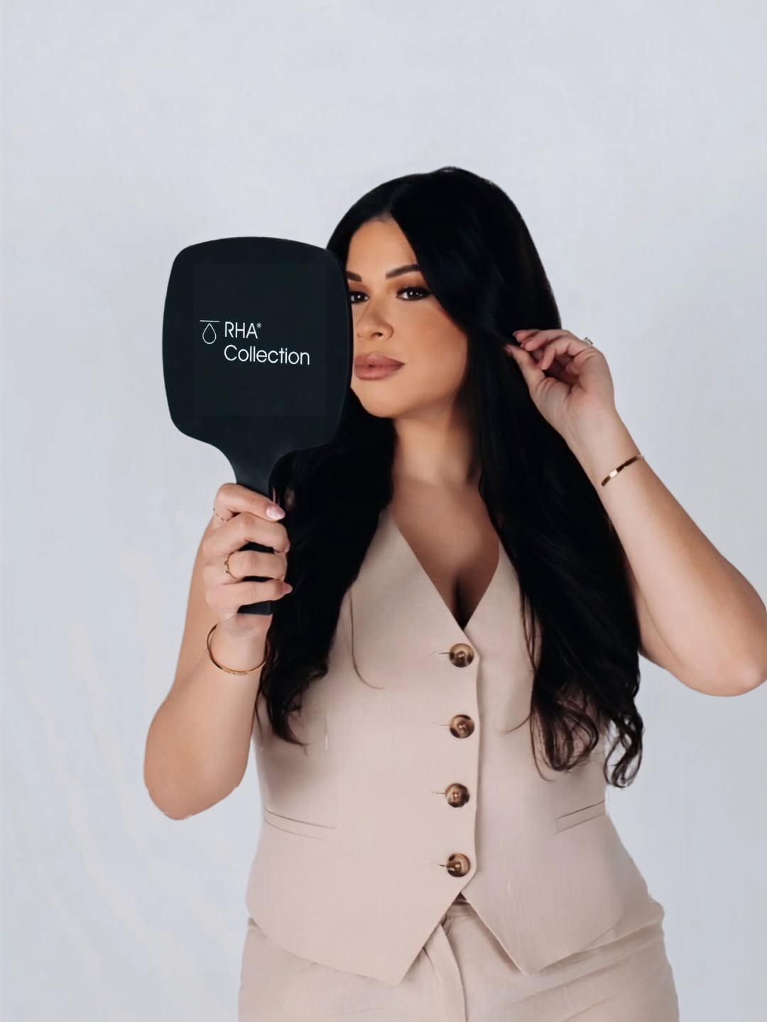 Woman with long dark hair in beige outfit holding a black hand mirror labeled RHA Collection.