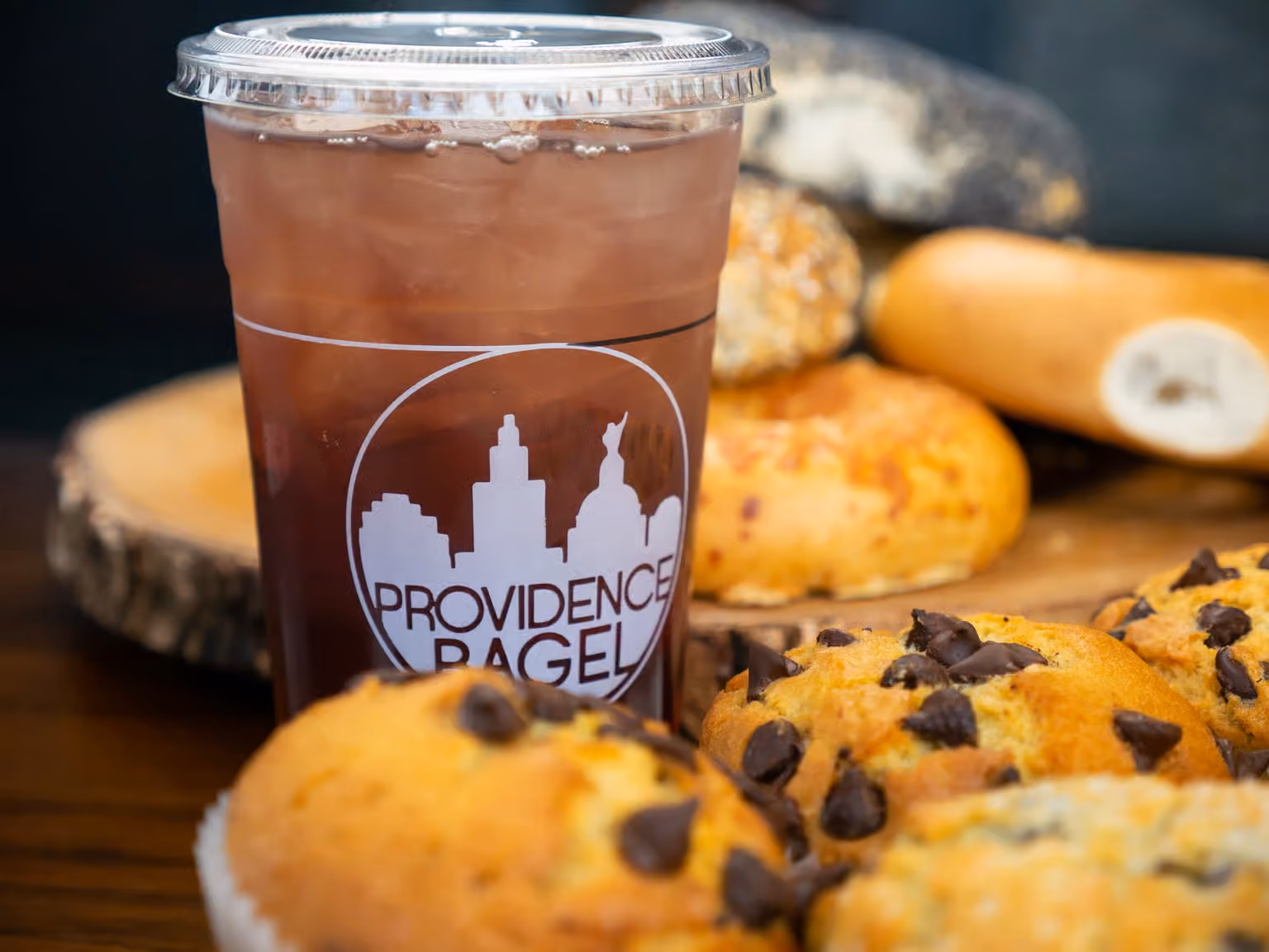 Iced drink in a Providence Bagel cup with chocolate chip muffins and bagels in the background.