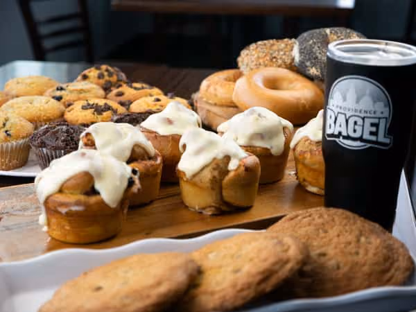 Variety of muffins, cookies, bagels, and a black cup with 'Bagel' logo on a wooden table.