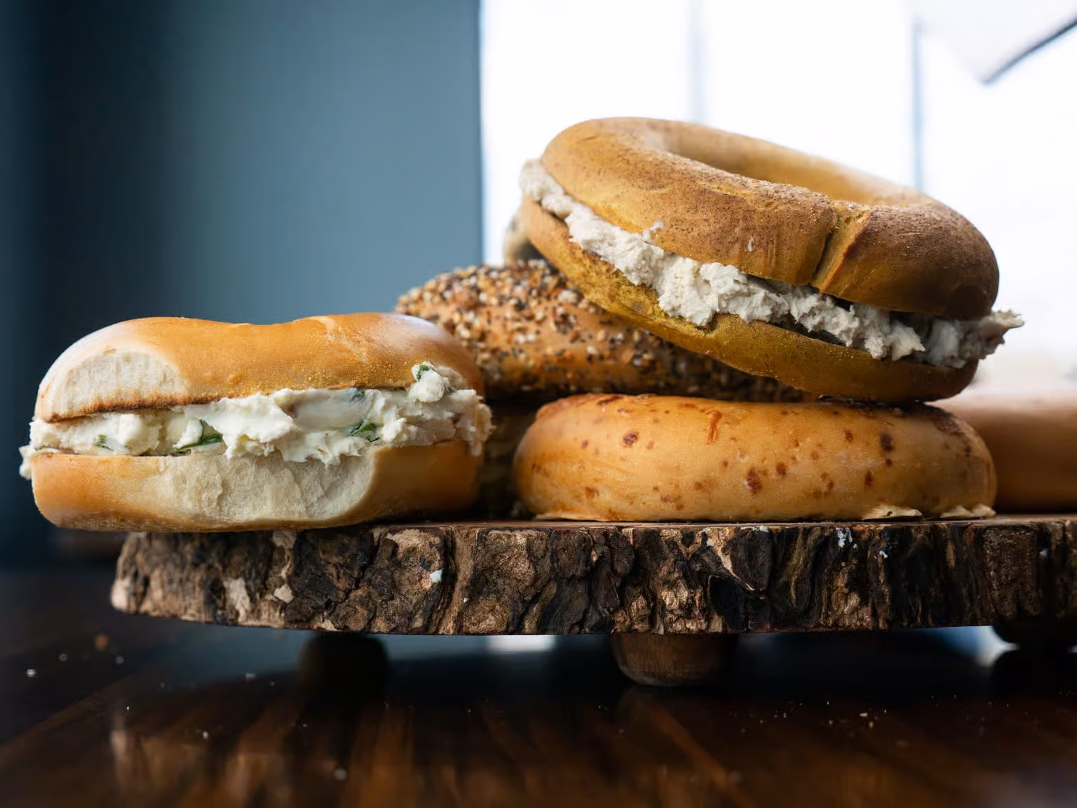 Four bagels with cream cheese on a rustic wooden serving board by a window.