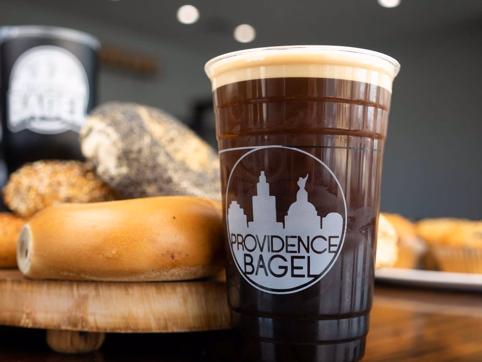 Plastic cup of dark coffee with foam on top and Providence Bagel logo, with assorted bagels in the background.