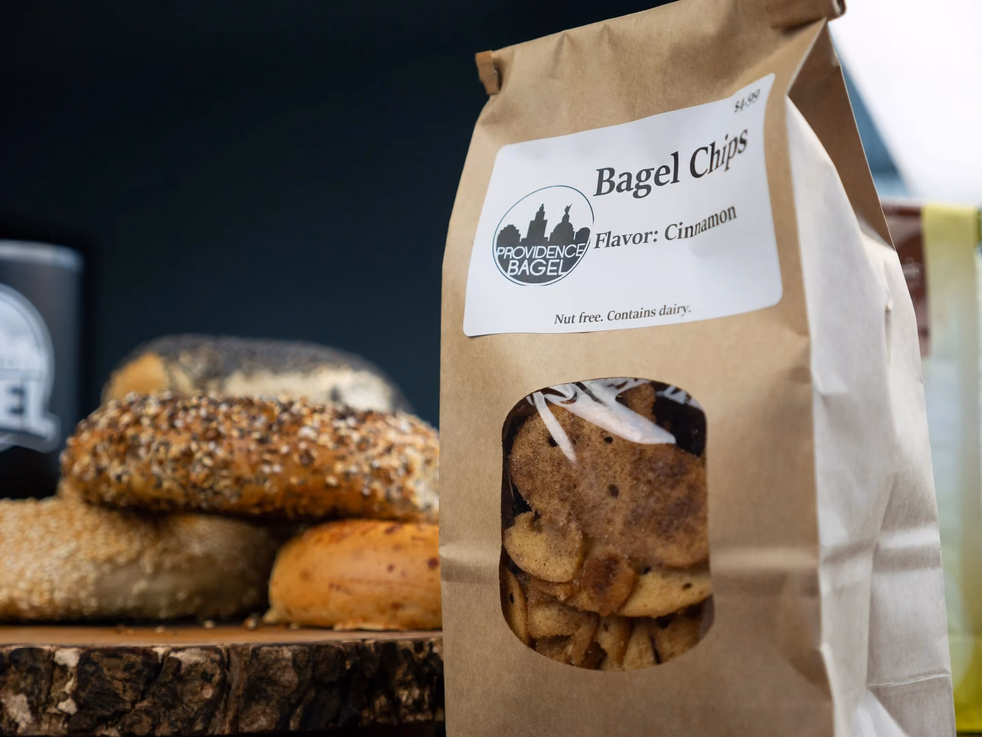 Bagel chips in a brown paper bag labeled cinnamon flavor with bagels in the blurred background.