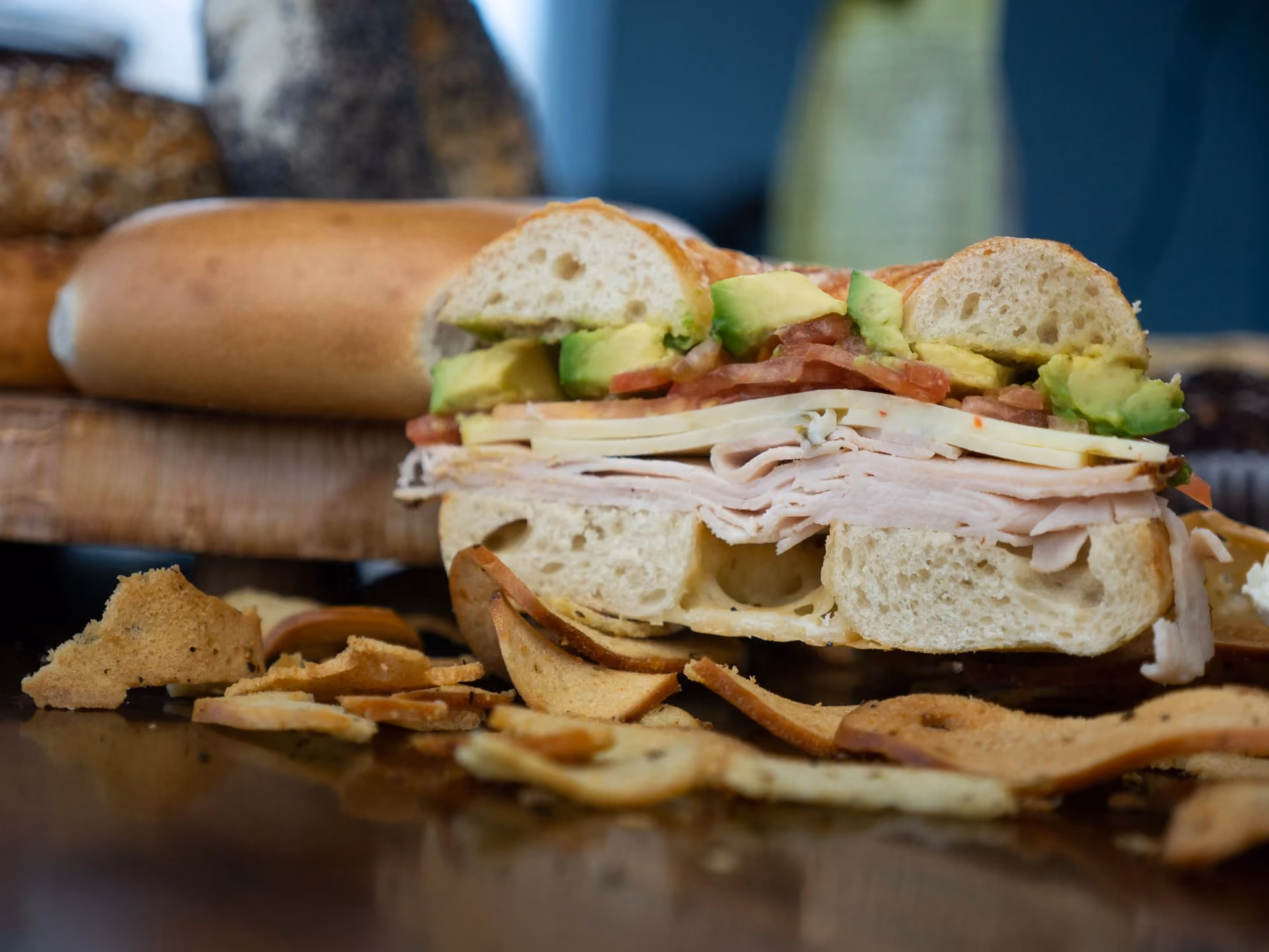 Turkey sandwich with avocado, tomato, and cheese on a bagel, served with potato chips.