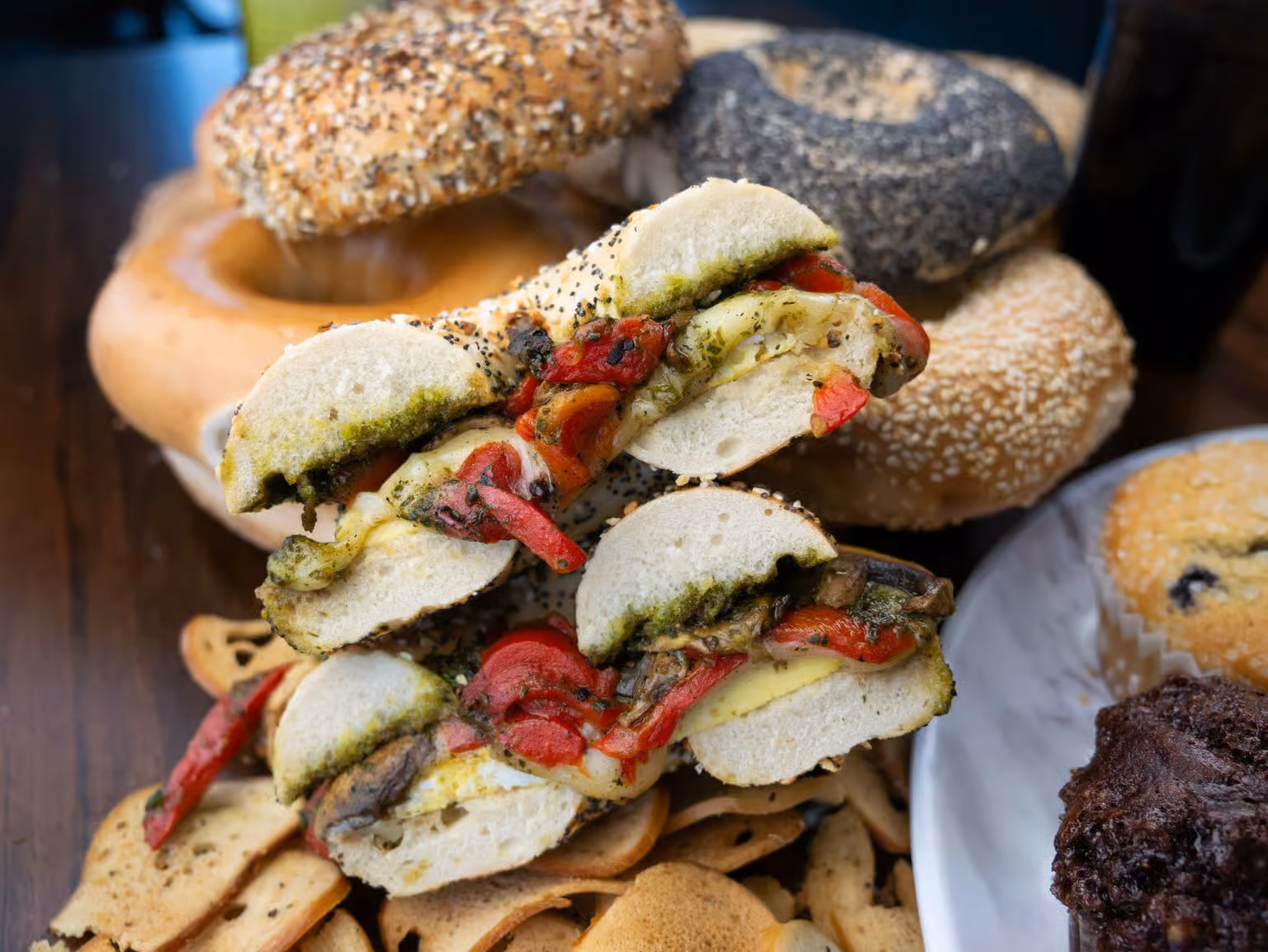 Stack of bagel halves with roasted red peppers and pesto, with assorted bagels and muffins in background.