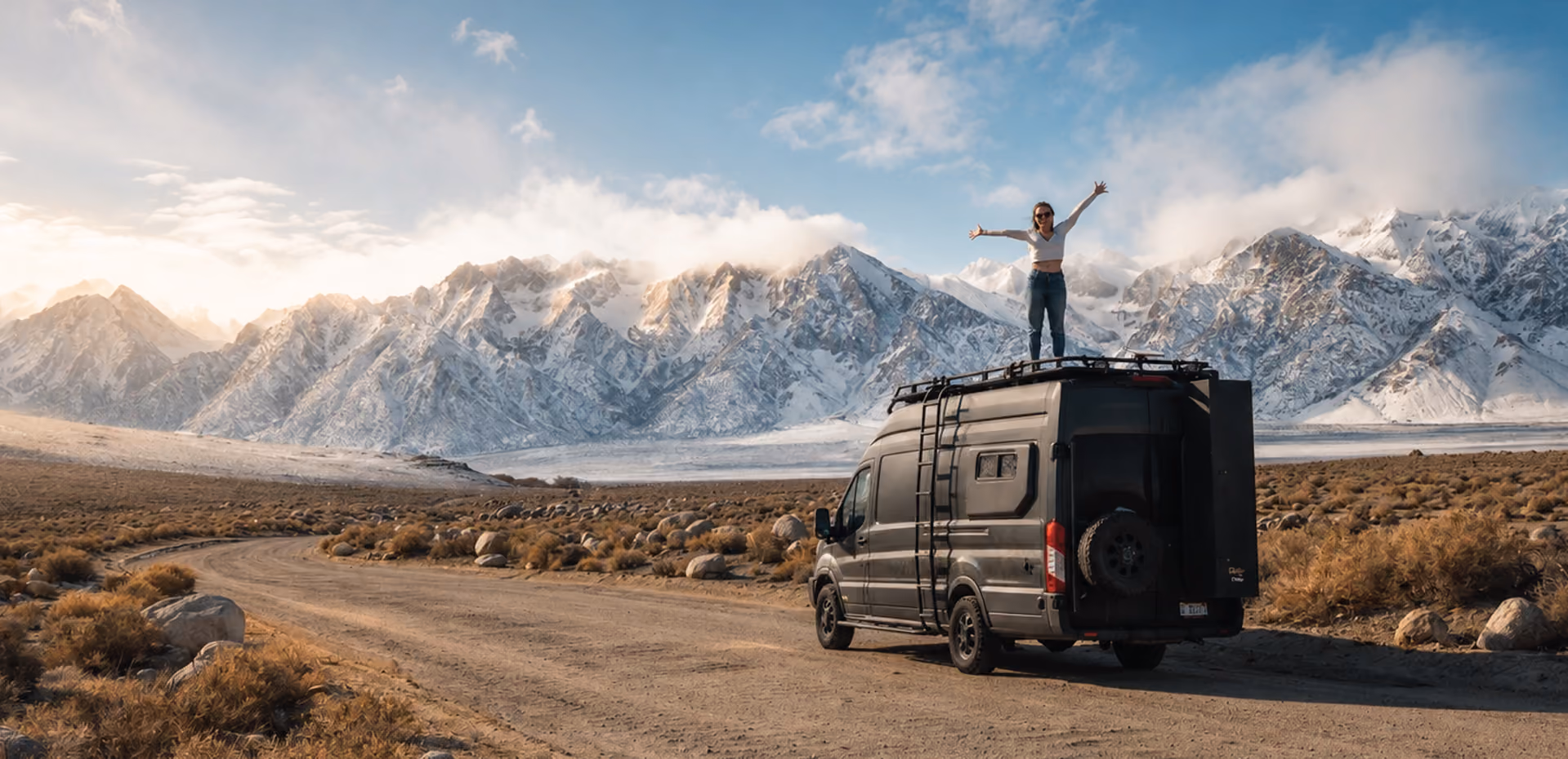 Owner standing arms spread on a Drifter campervan roof before snow-capped mountains